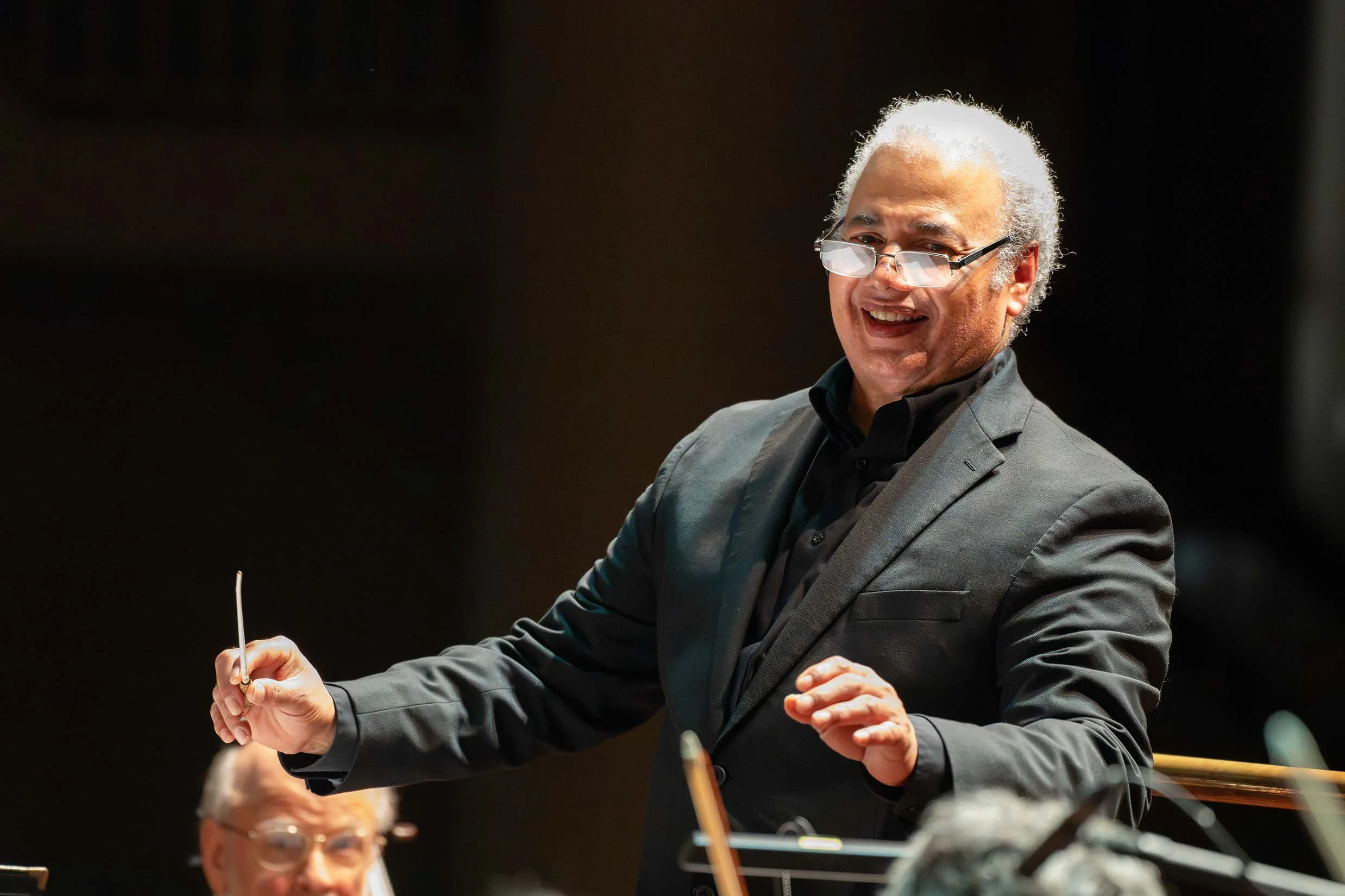 A man in a black suit and glasses conducting an orchestra, with a person in the background playing a string instrument.