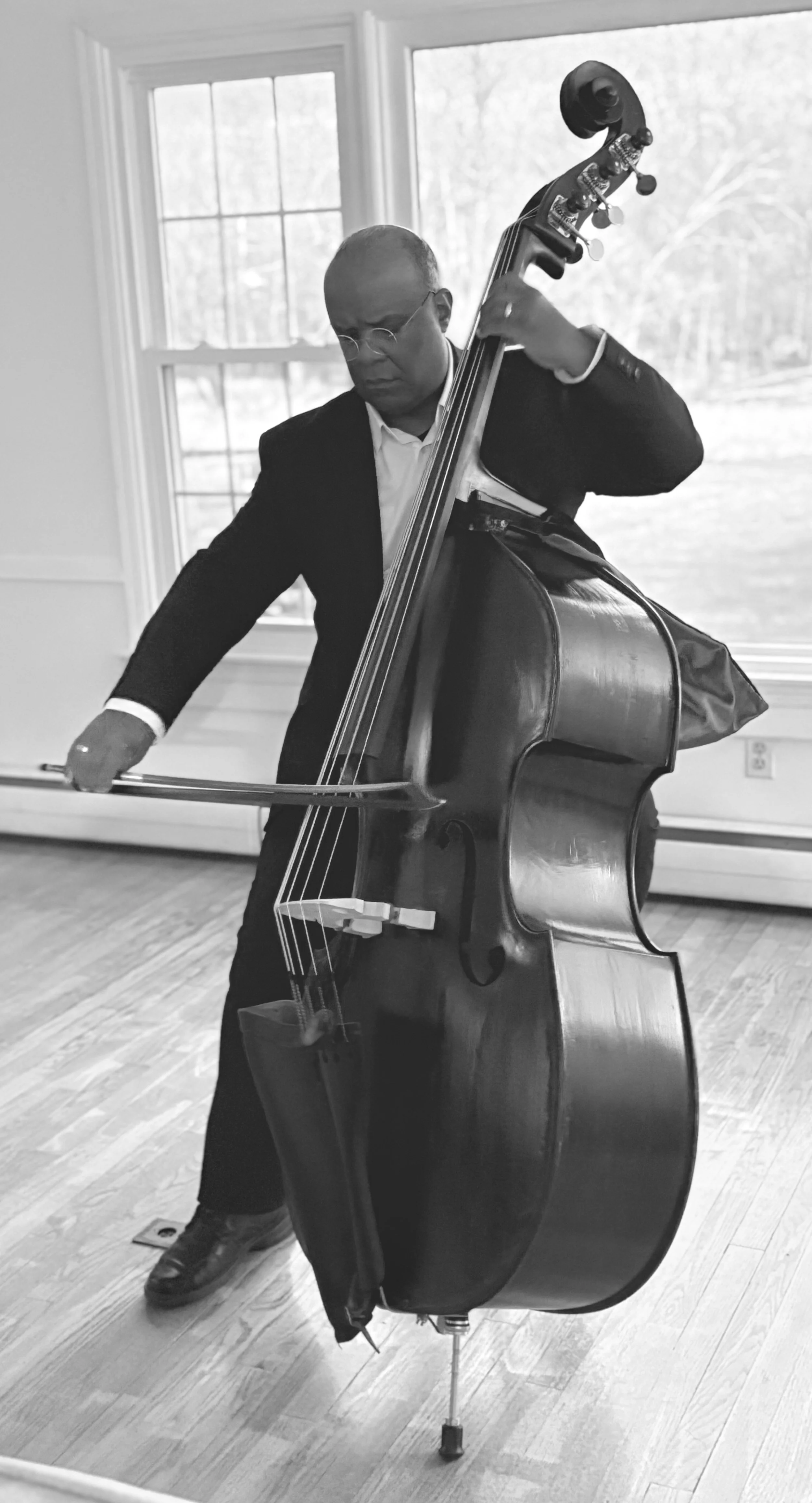 A man wearing glasses and a suit is playing a double bass indoors near a window, with the room having hardwood floors.