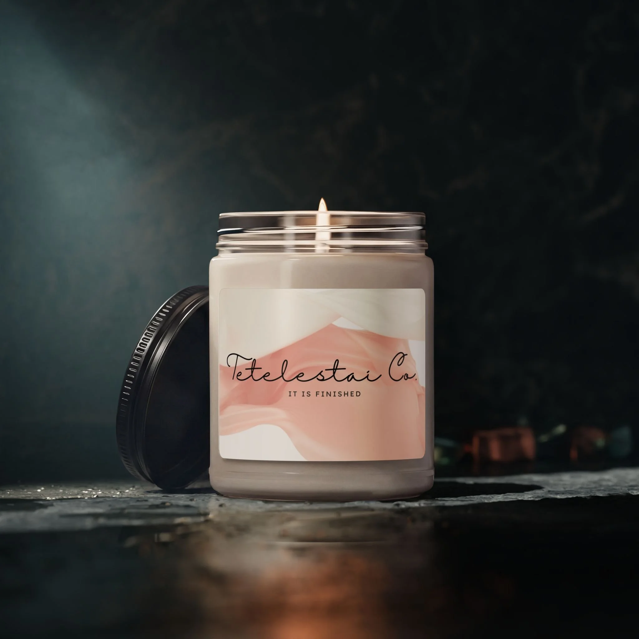 A candle with a small flame on a dark surface with a blurred background featuring the brand label for Tetelestai Co..