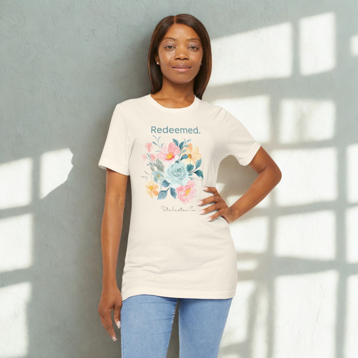 A woman wearing a beige T-shirt with a floral design and the word 'Redeemed.' standing against a light-colored wall with sunlight shadows.