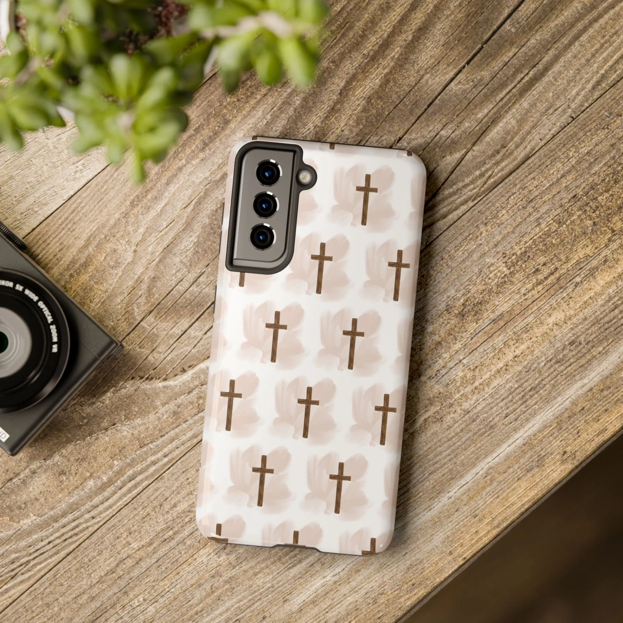 A smartphone with a white patterned case featuring cross-shaped designs, resting on a wooden surface beside a plant and a camera.