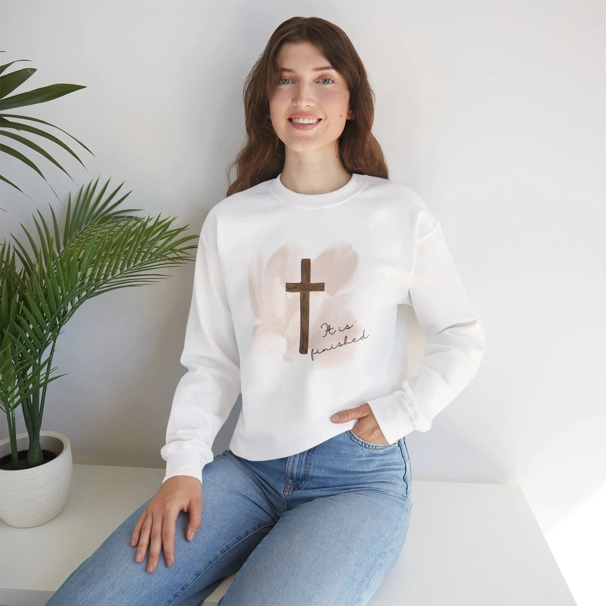 A woman wearing a white sweatshirt with a cross and the words "It is finished" stands next to a white vase with a green plant, against a plain white background.