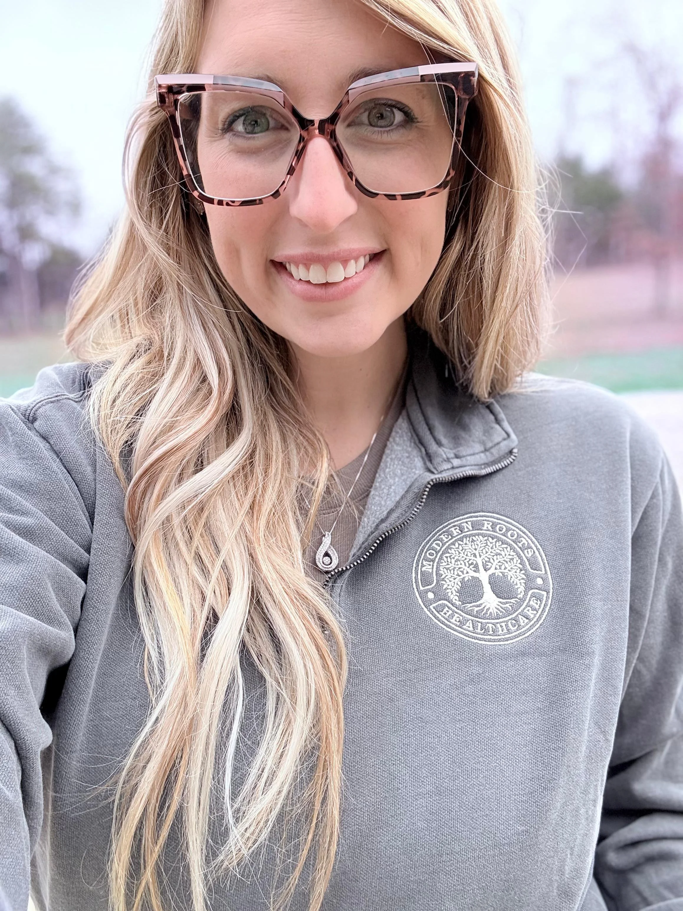 Abby Carter with long blonde hair wearing glasses with a leopard print frame, smiling. She is wearing a gray jacket with a logo on the chest.