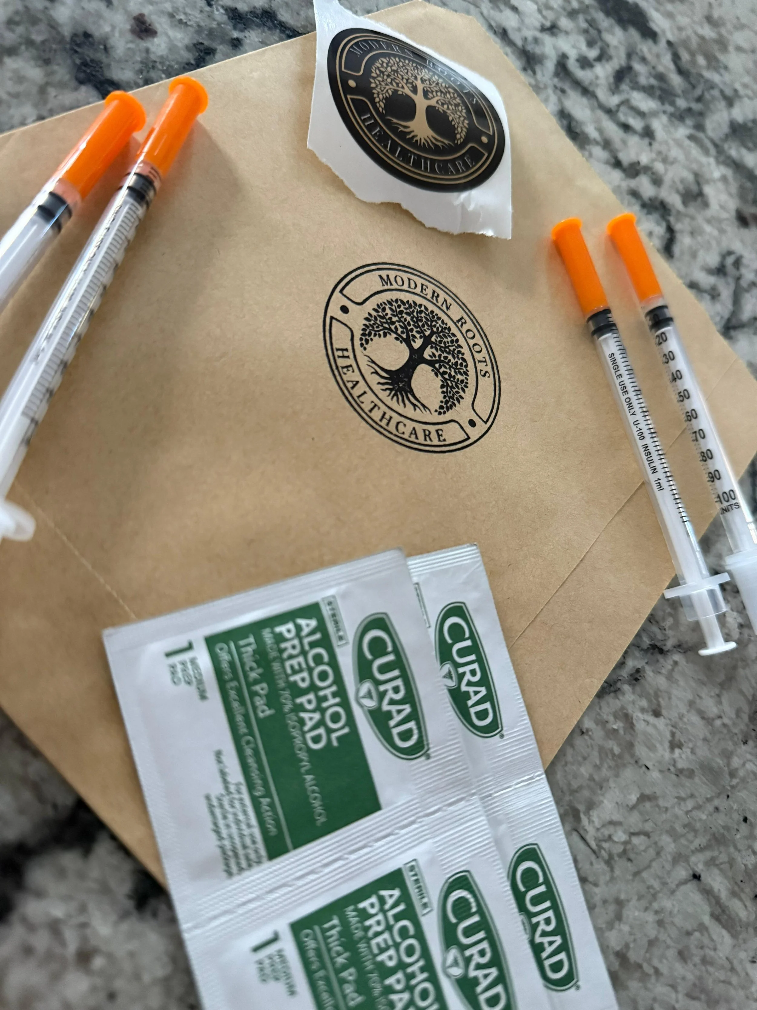 A brown paper bag with a black logo of a tree surrounded by the words "Modern Roots Healthcare." Four orange-lid syringes and several green and white tea packets are on the bag. The background is a gray granite countertop. | Modern roots healthcare