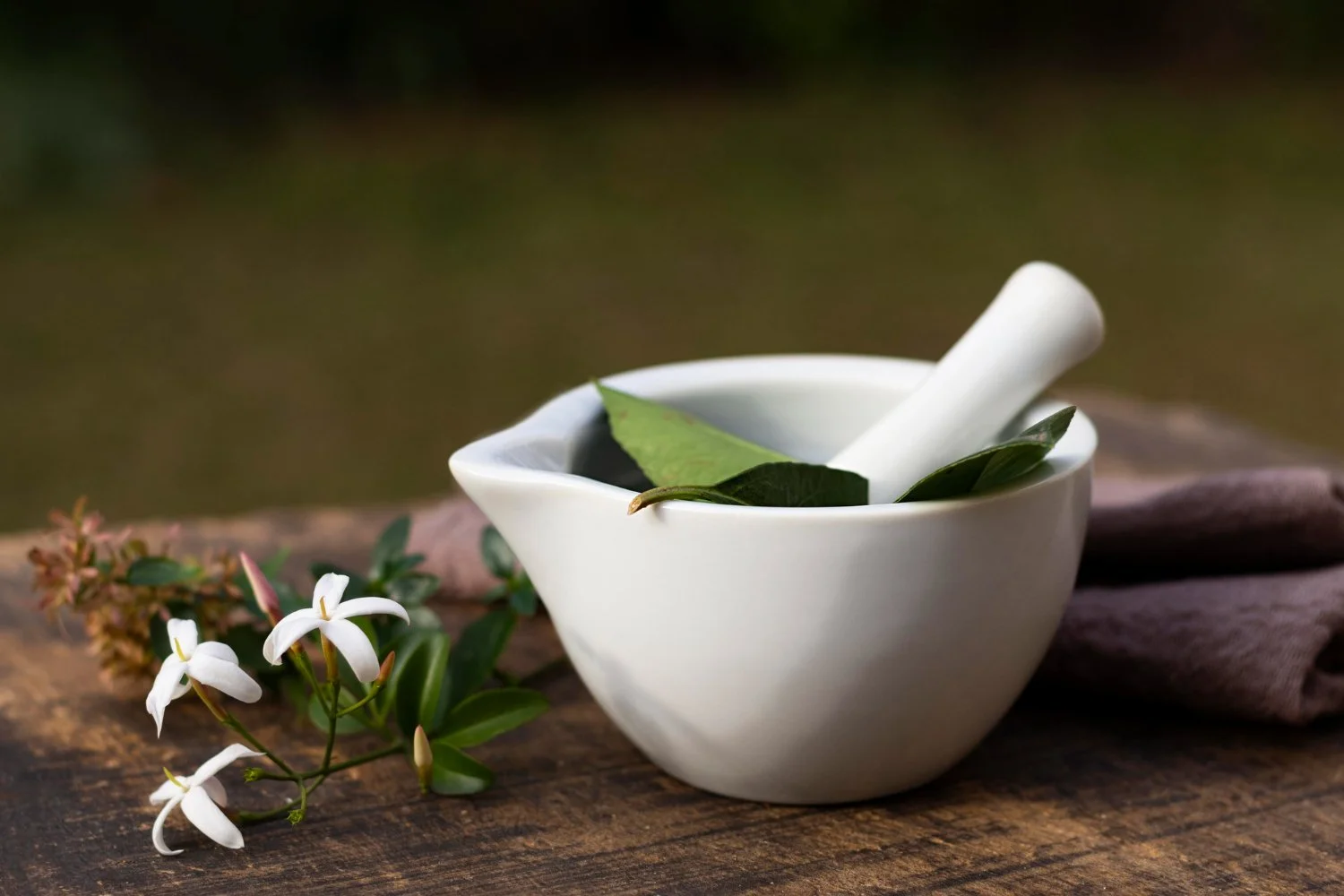 Modern roots healthcare  | A white ceramic mortar and pestle with green leaves inside, placed on a rustic wooden surface with white flowers and green foliage beside it.