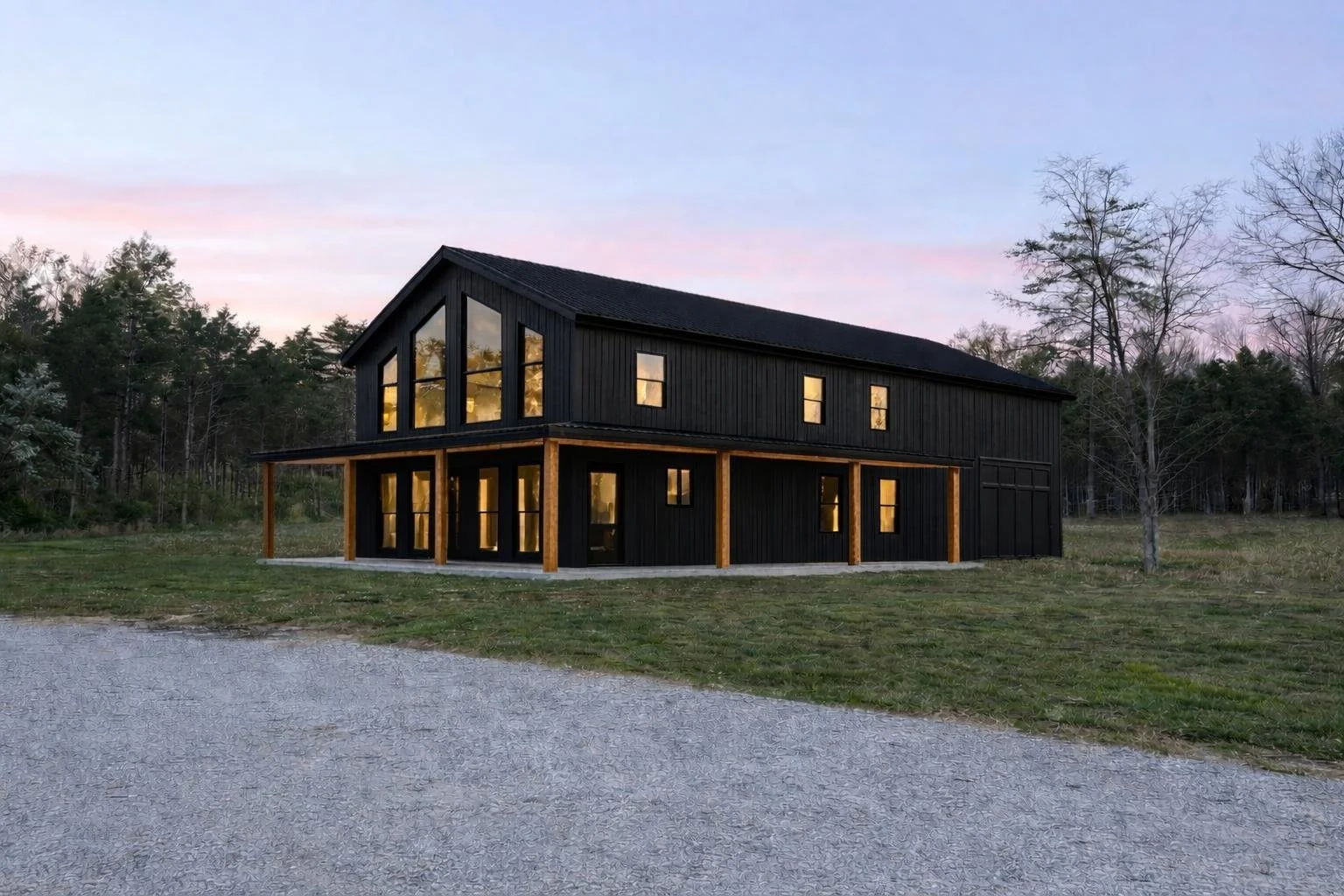 Modern two-story black house with large windows and wooden accents, set on a grassy field with trees and a gravel driveway at sunset.