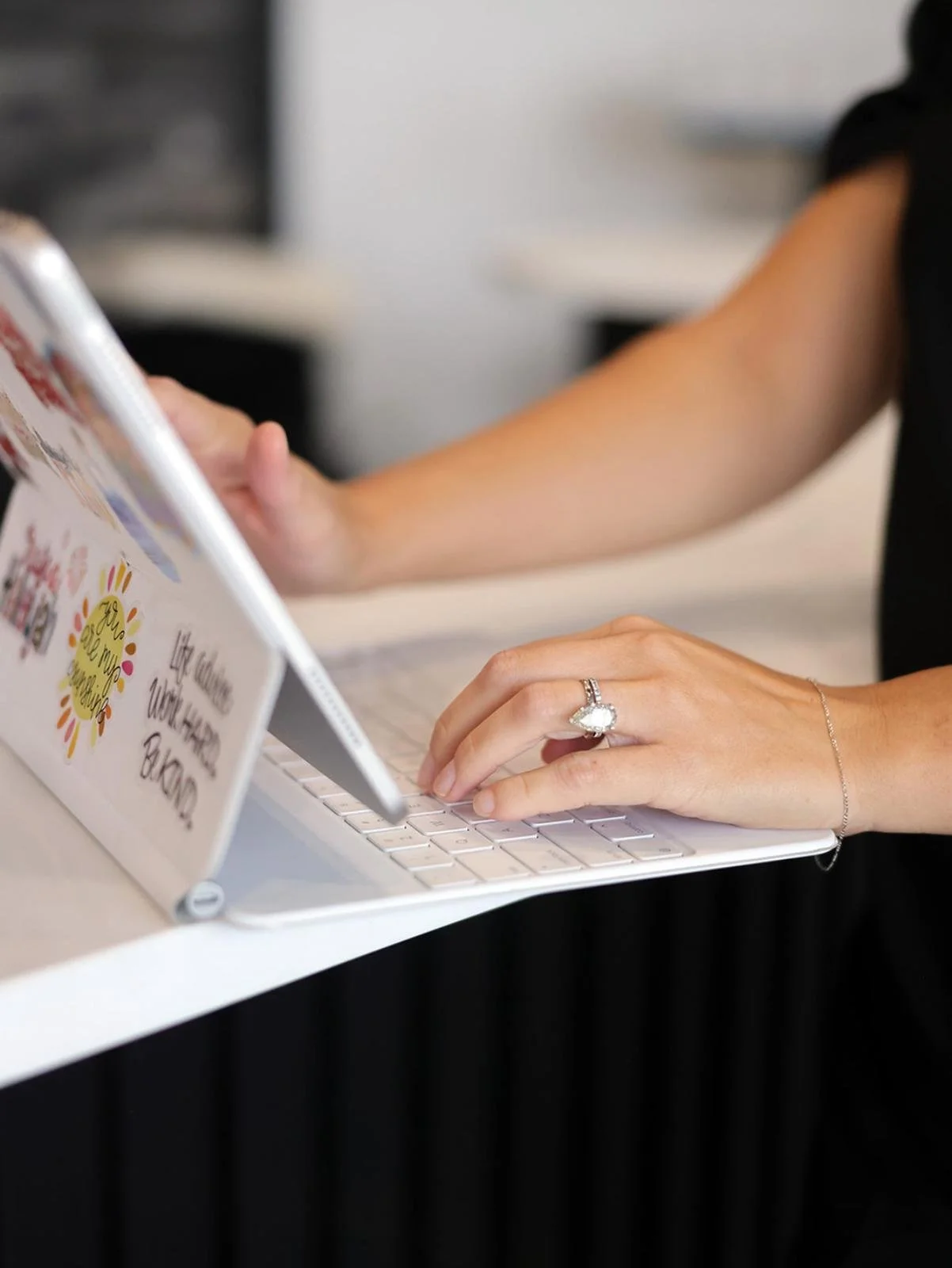 Abby Carter hand with an engagement ring and bracelet typing on a white laptop keyboard, with a woman holding a flyer or brochure in the background.