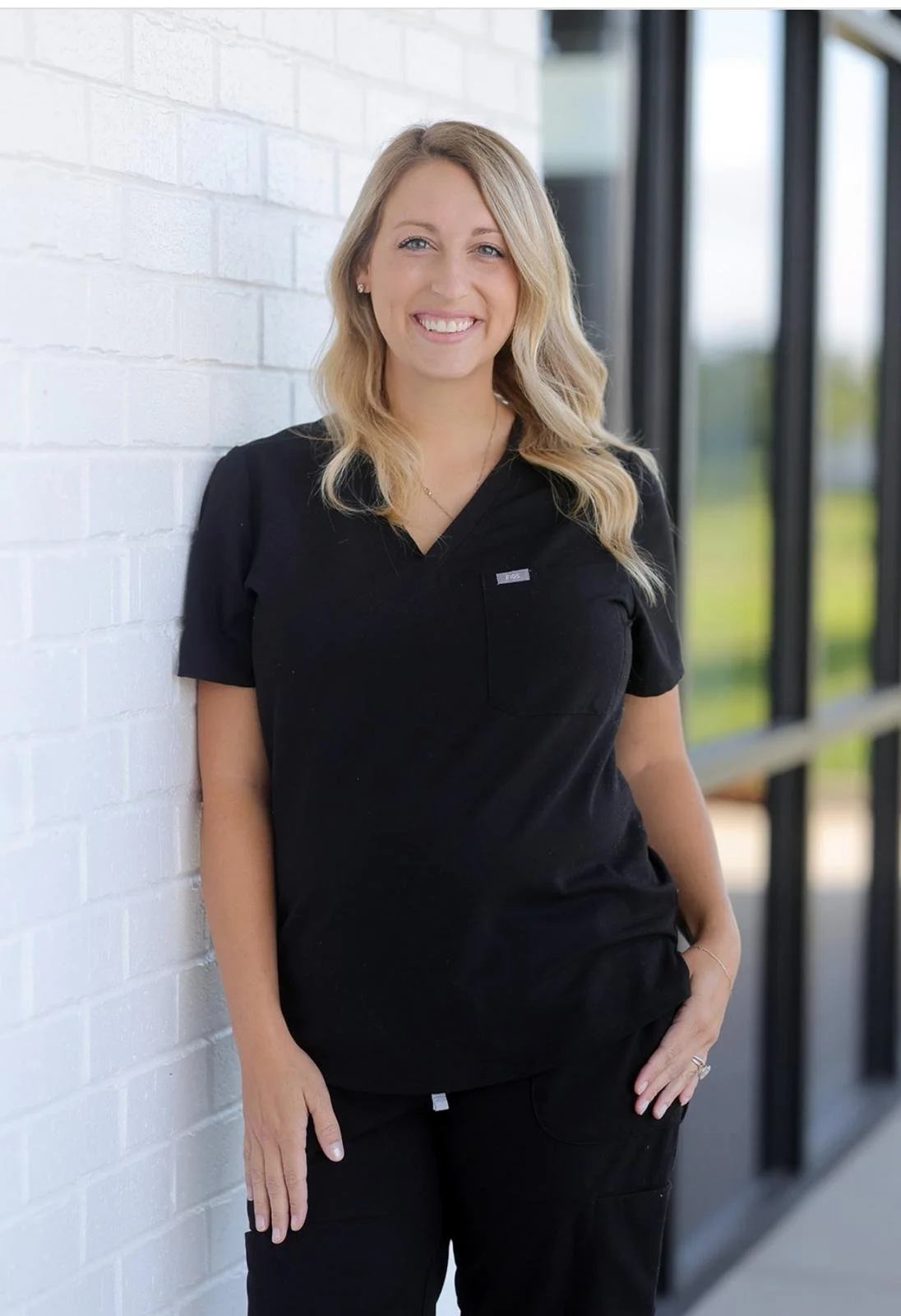 Abby Carter smiling woman with blonde hair wearing black scrubs, standing against a white brick wall near large windows.