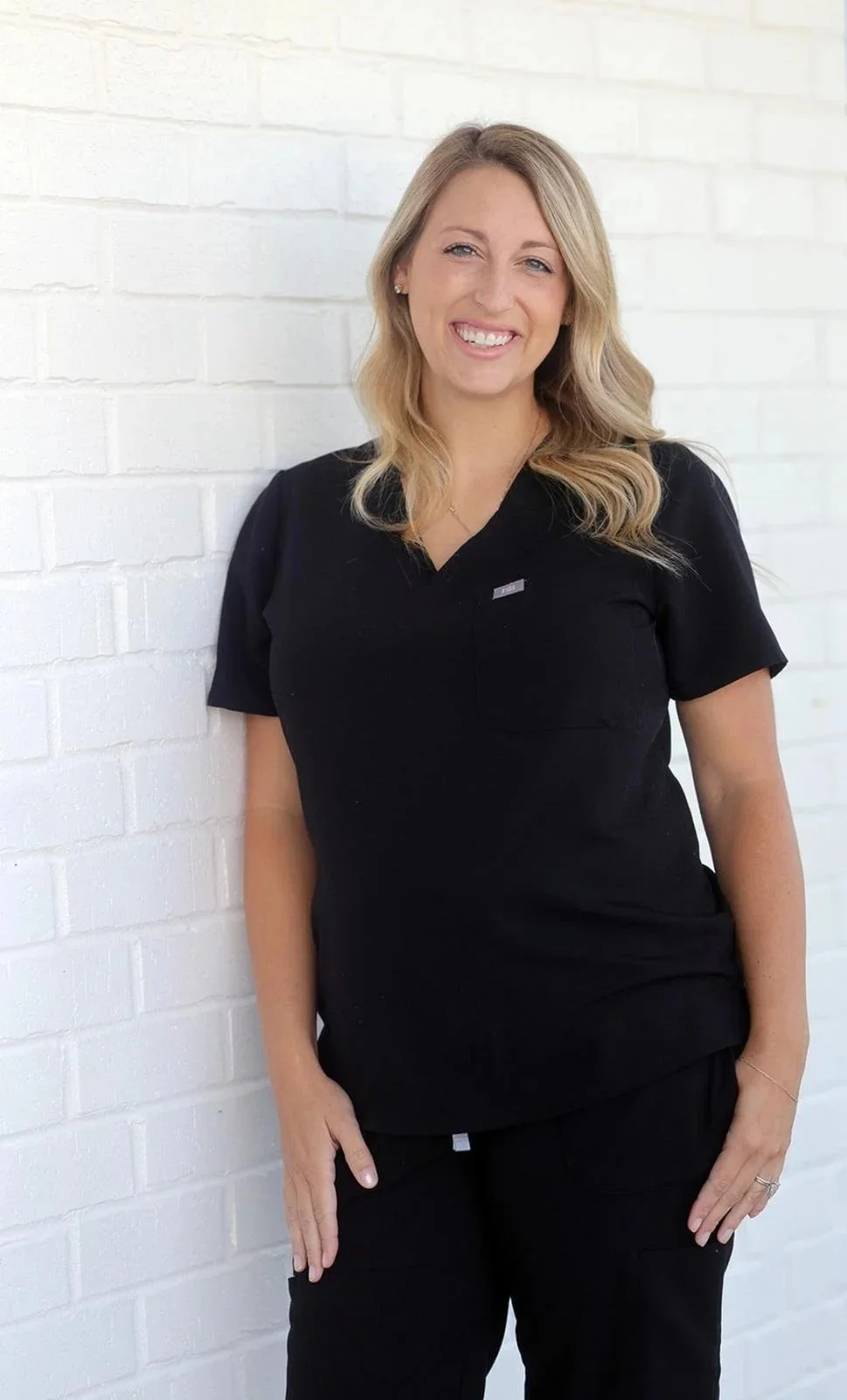 Abby Carter in black scrubs standing against a white brick wall, smiling.