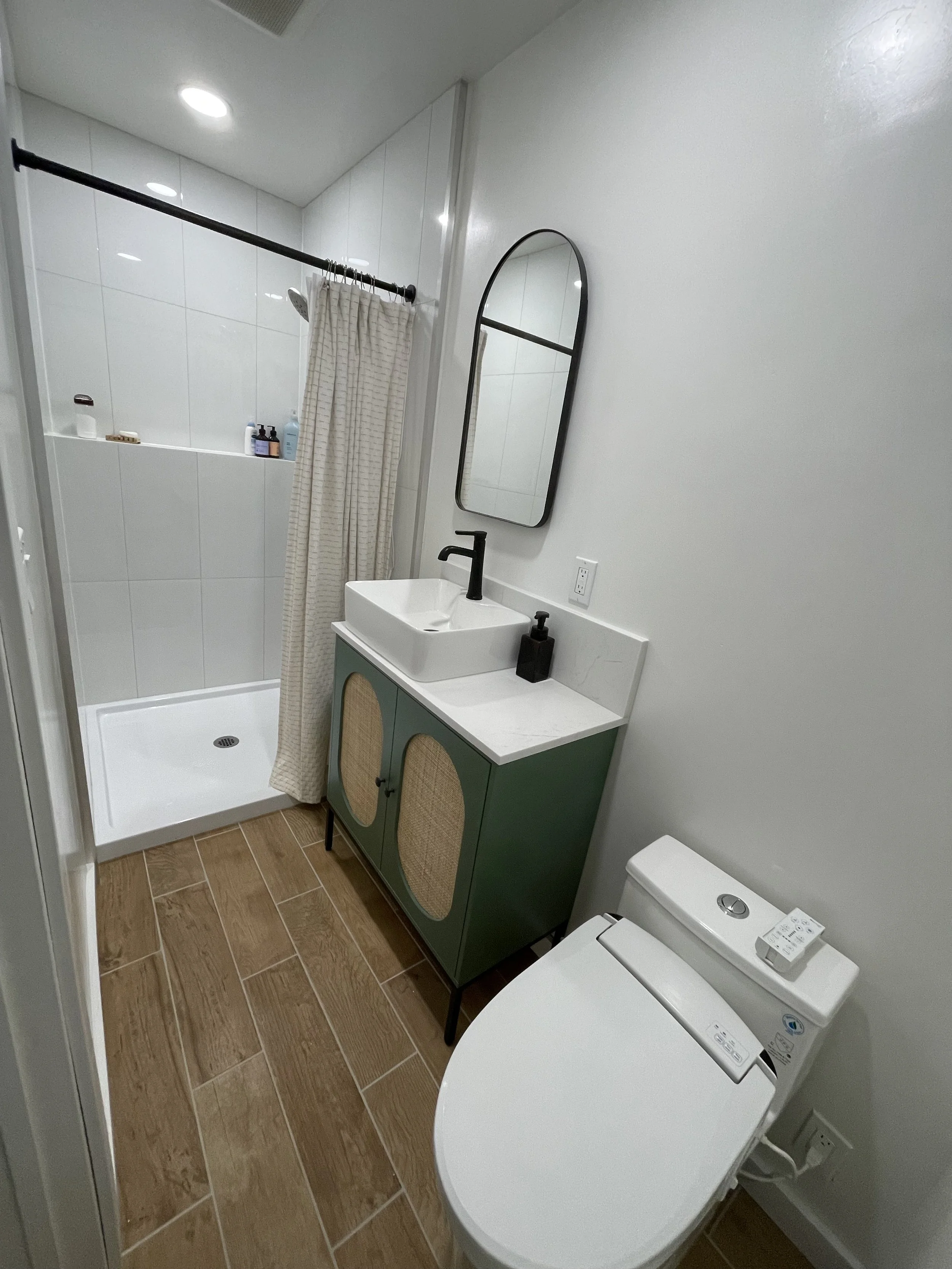 Modern bathroom with a shower area enclosed by a curtain, a sink with a black faucet, a mirror, a washing machine, and a toilet, all with wood-tone tile flooring and white walls.
