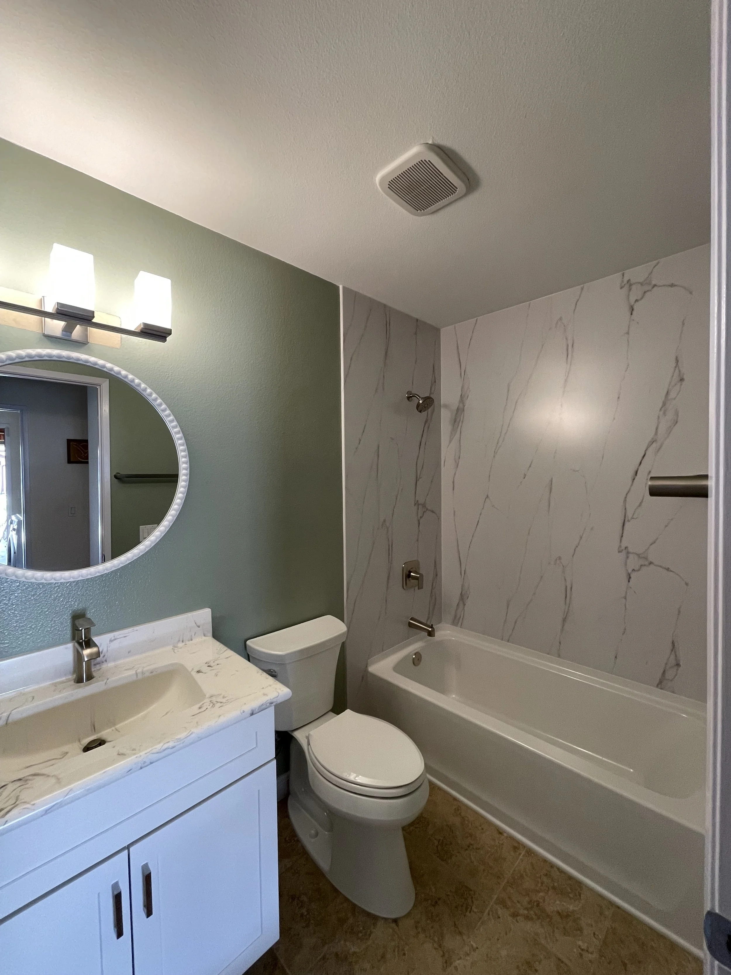 Bathroom with a white vanity with two sinks, a mirror with lights, a white toilet, a bathtub with marble-patterned walls, and a vent on the ceiling.