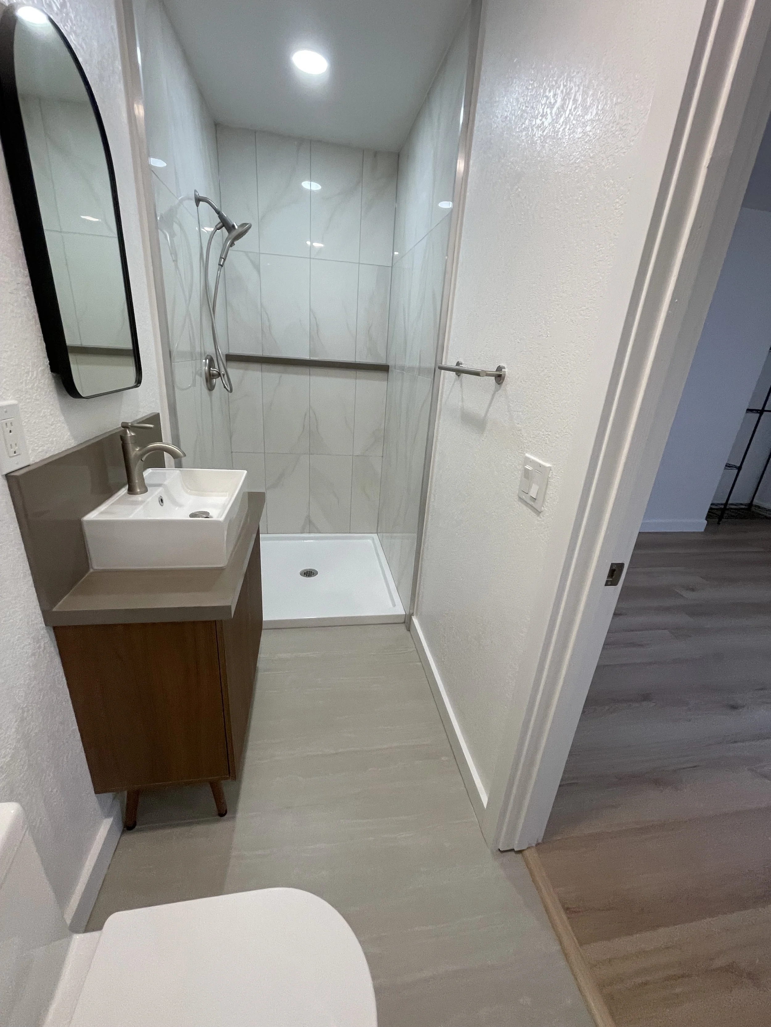 A small bathroom with a shower stall that has a clear glass door, a wall-mounted showerhead, a small rectangular sink with a bronze faucet, a mirror mounted vertically on the wall, a beige vanity, a towel bar on the wall, a toilet partially visible i