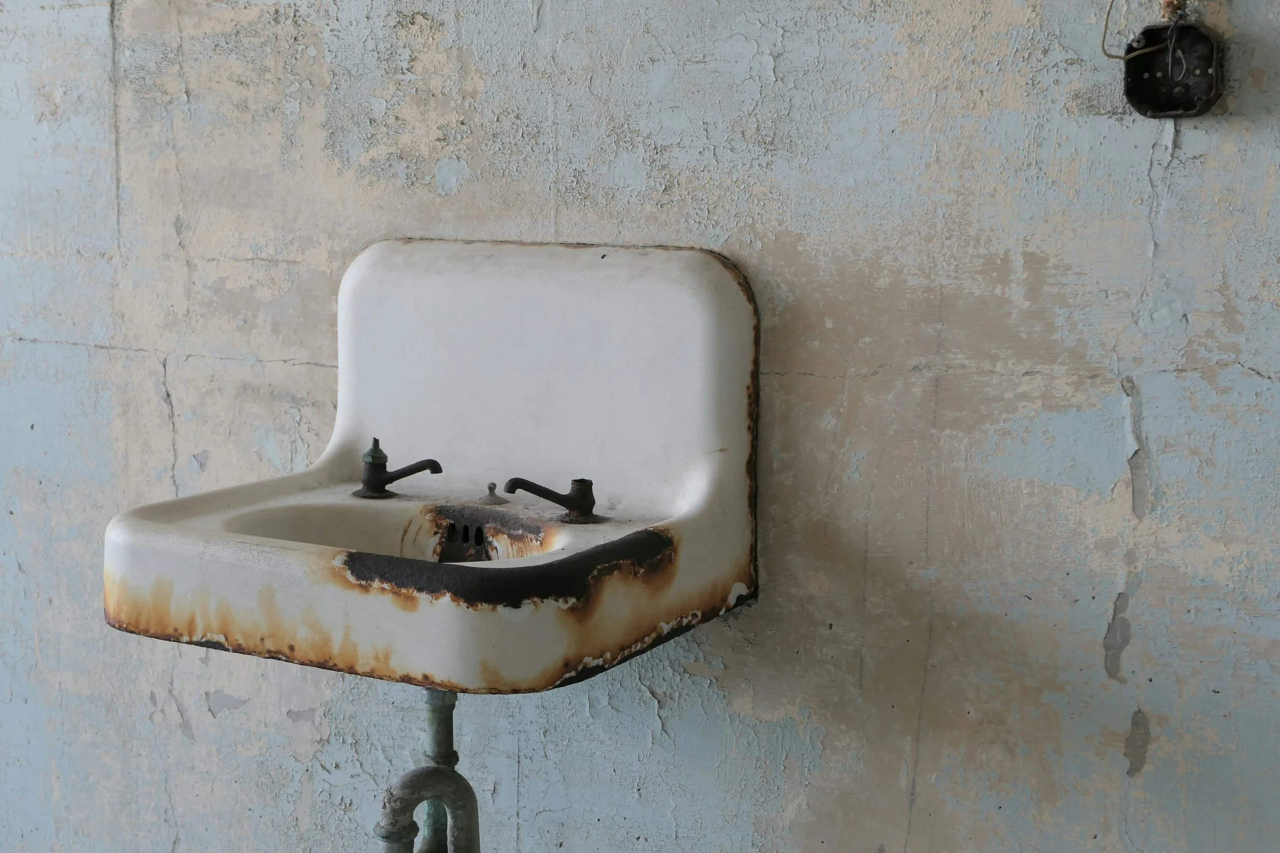 An old, rusty wall-mounted sink with a blackened drain, set against a peeling, textured wall. An electrical outlet is visible nearby.