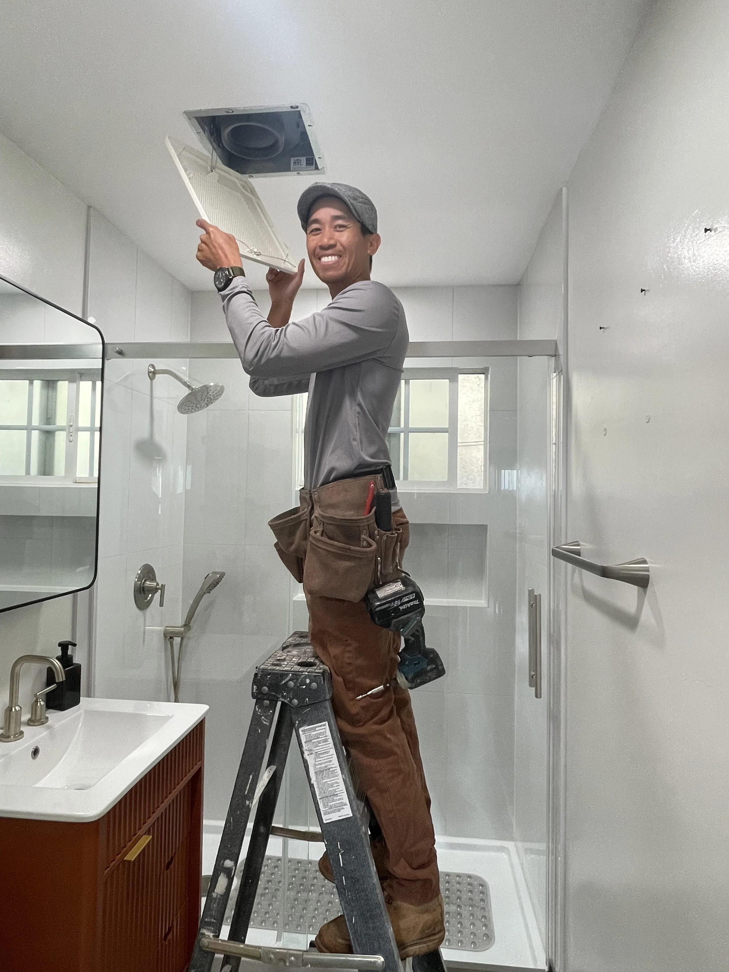 A man wearing a gray cap, gray shirt, and brown work pants stands on a step ladder in a bathroom, opening an exhaust fan on the ceiling. He has a tool belt and a drill attached to his pants and is smiling at the camera.