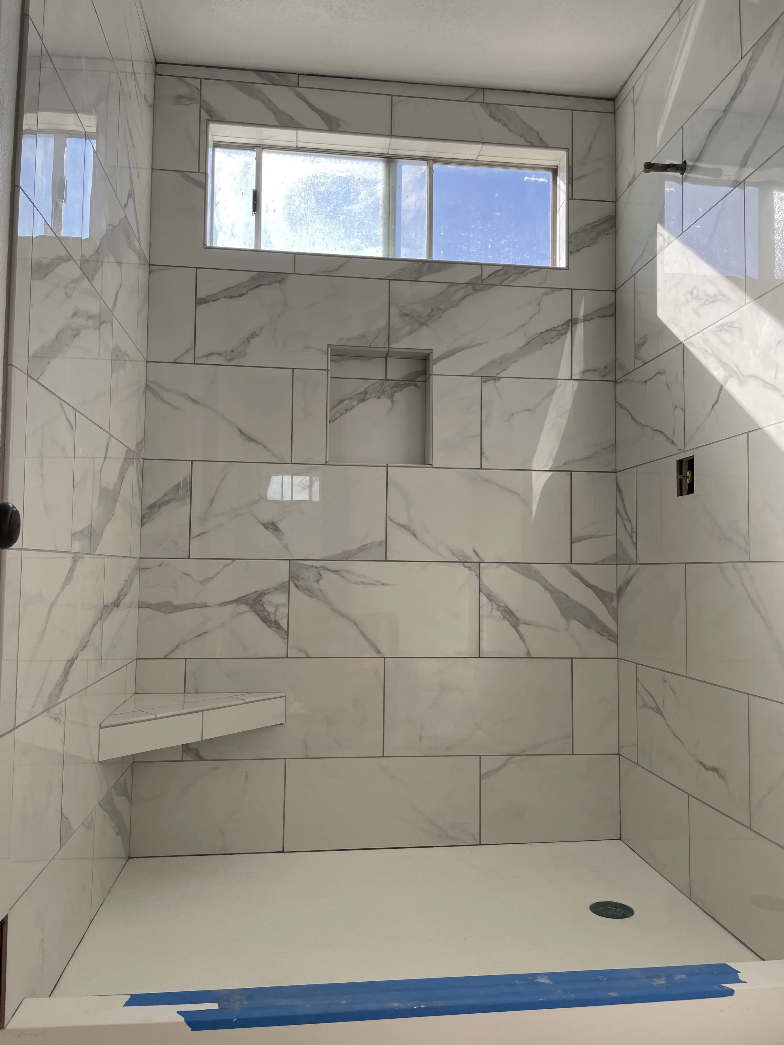 A modern shower with marble tiles on the walls, a small built-in wall shelf, a window at the top, and a drain on the floor.