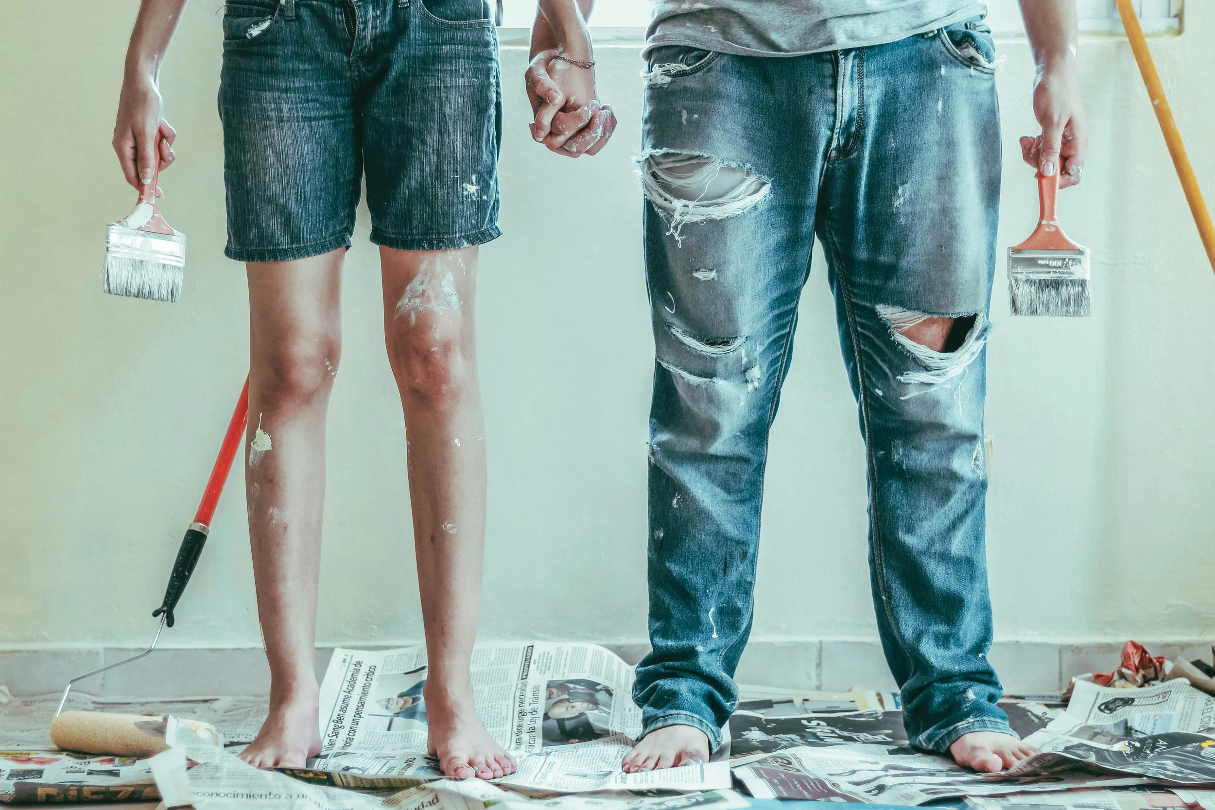 Two people standing side by side, holding hands, with paint on their legs and clothes. They are standing on a floor covered with scattered newspapers. One person is barefoot, while the other is wearing jeans. They are holding paintbrushes and a roller, indicating painting activity.
