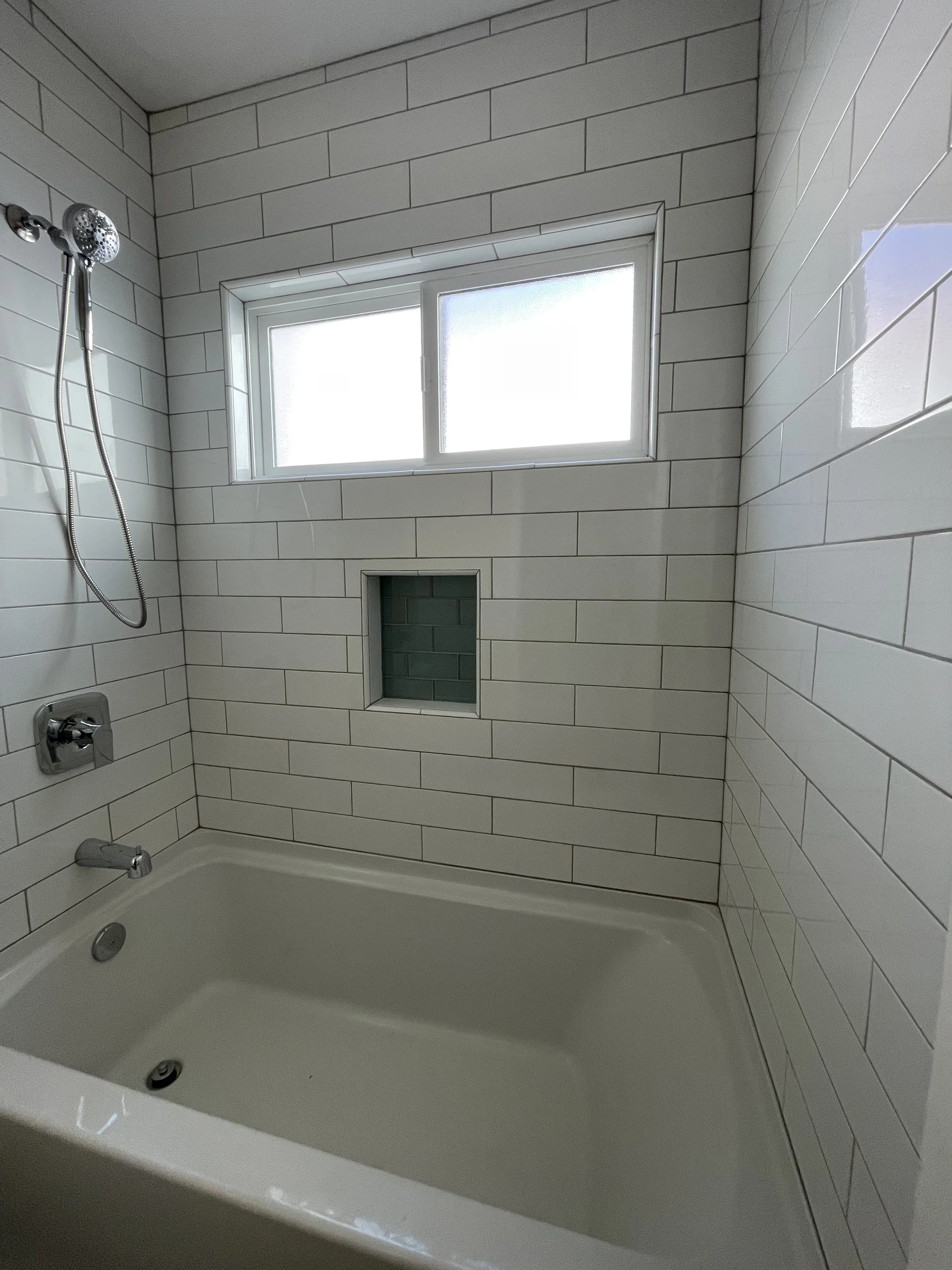 A bathroom with white subway tile walls, a window, a built-in niche, and a bathtub with a showerhead.