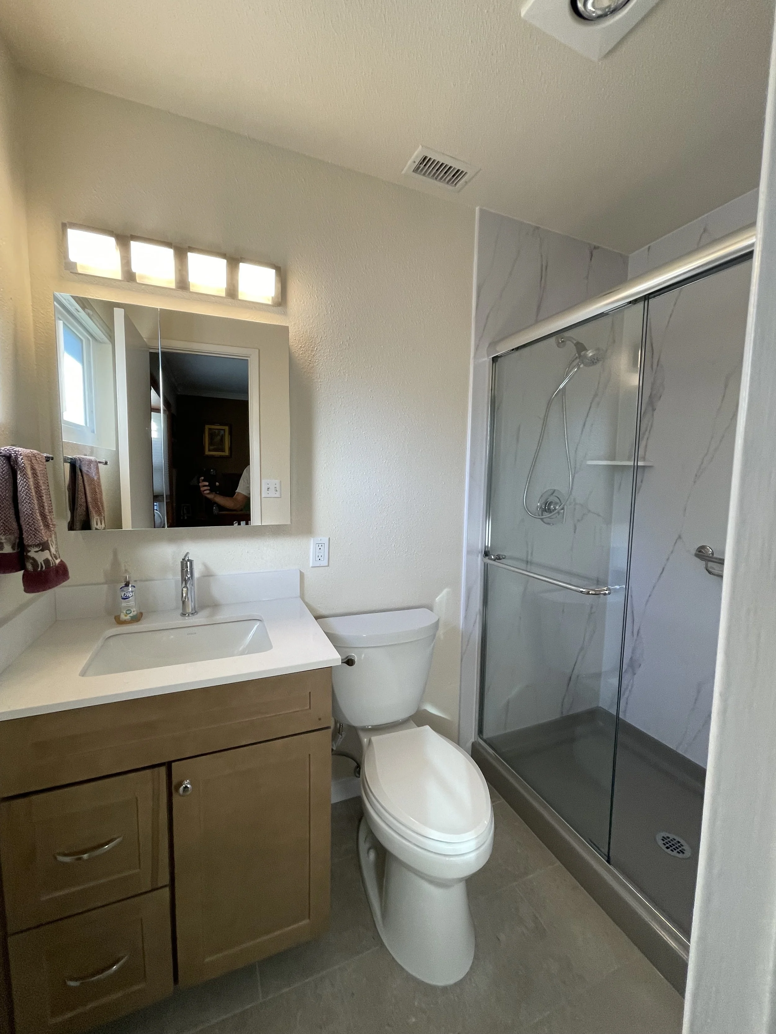 A bathroom with a vanity mirror, a white sink, a wooden cabinet, a toilet, and a glass shower enclosure with marble tiles. There is a window and a towel rack with towels.