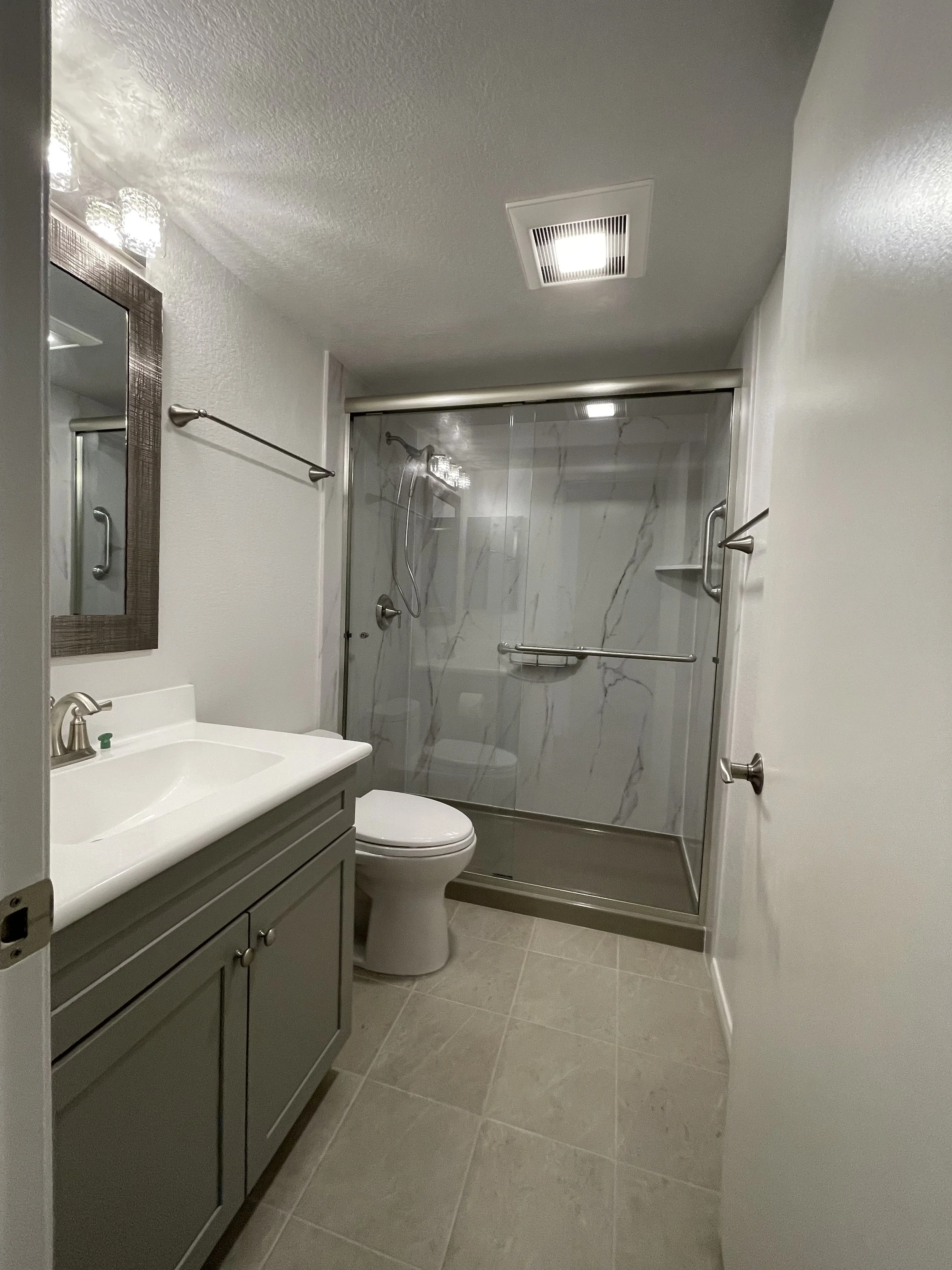 Bathroom with gray vanity, mirror, toilet, walk-in shower with marble walls, and beige tiled floor.