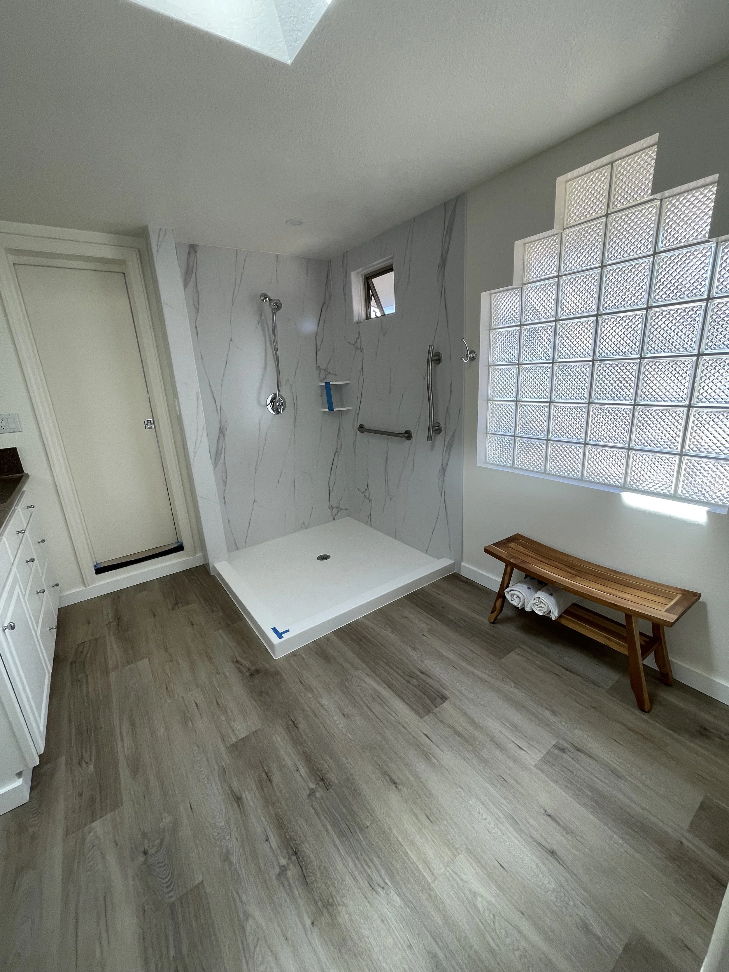 Bathroom with walk-in shower featuring marble walls, a small window, and glass block wall. There is a wooden bench with rolled white towels, and wood-look flooring.