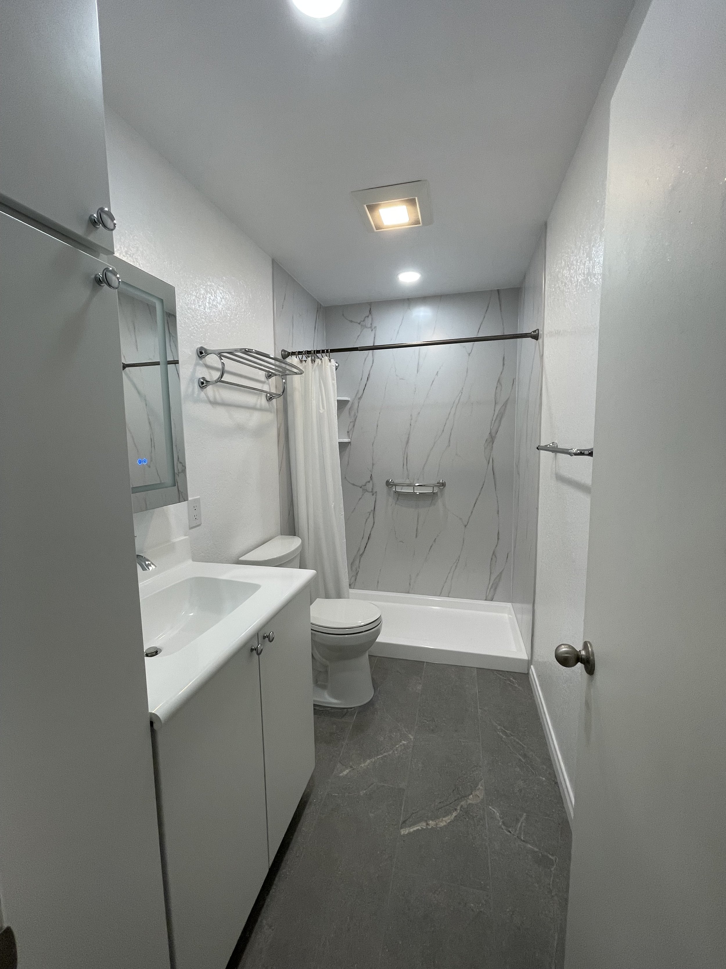 A small bathroom with a gray tile floor, white walls, a shower with a curtain and metal rod, a toilet, a small white sink with cabinet, a mirror, a towel rack, and modern ceiling lights.