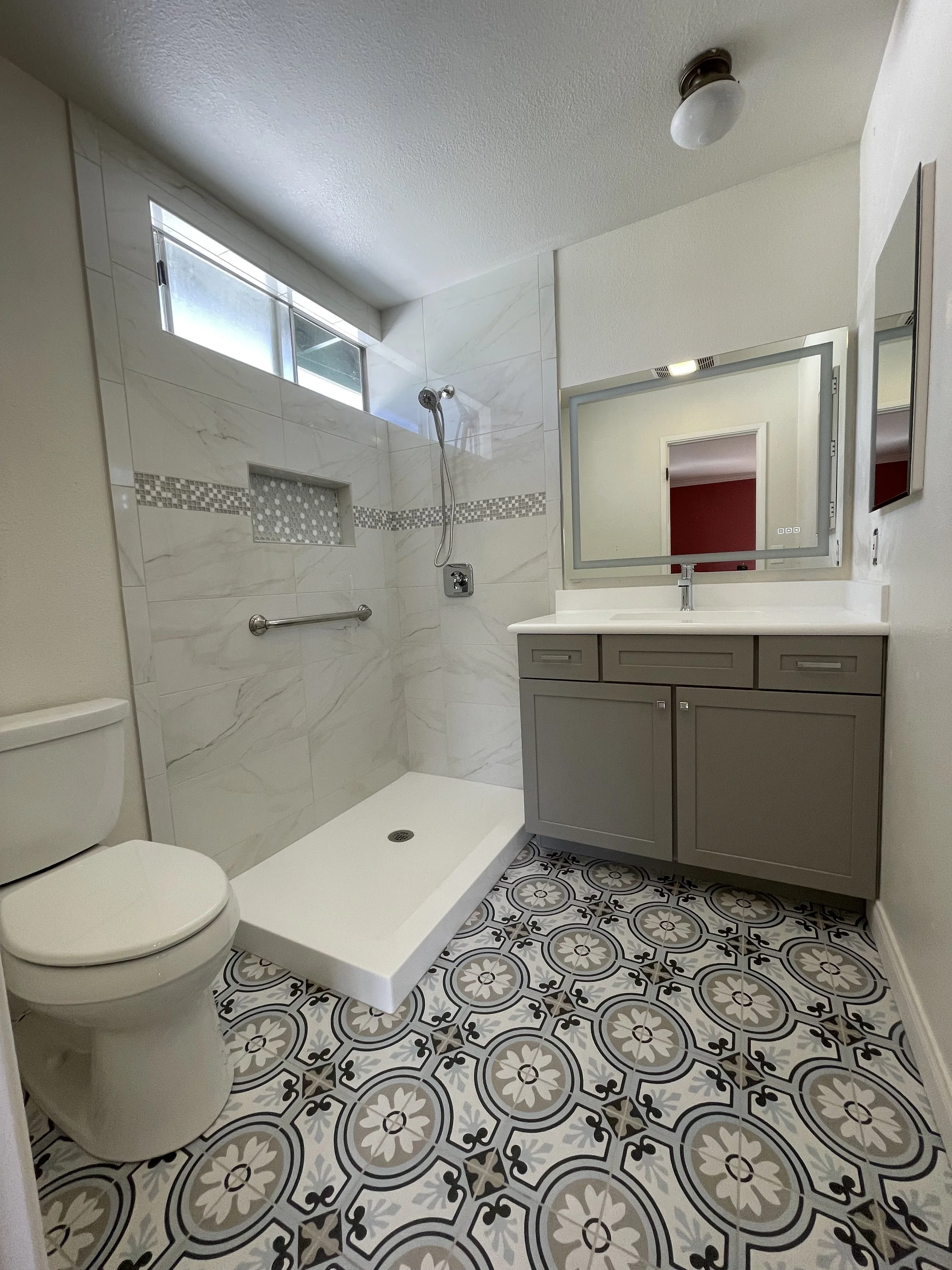 A modern bathroom with patterned tile flooring, a toilet, a shower with white marble tiles and a small window, a gray vanity with a mirror, and a ceiling light fixture.