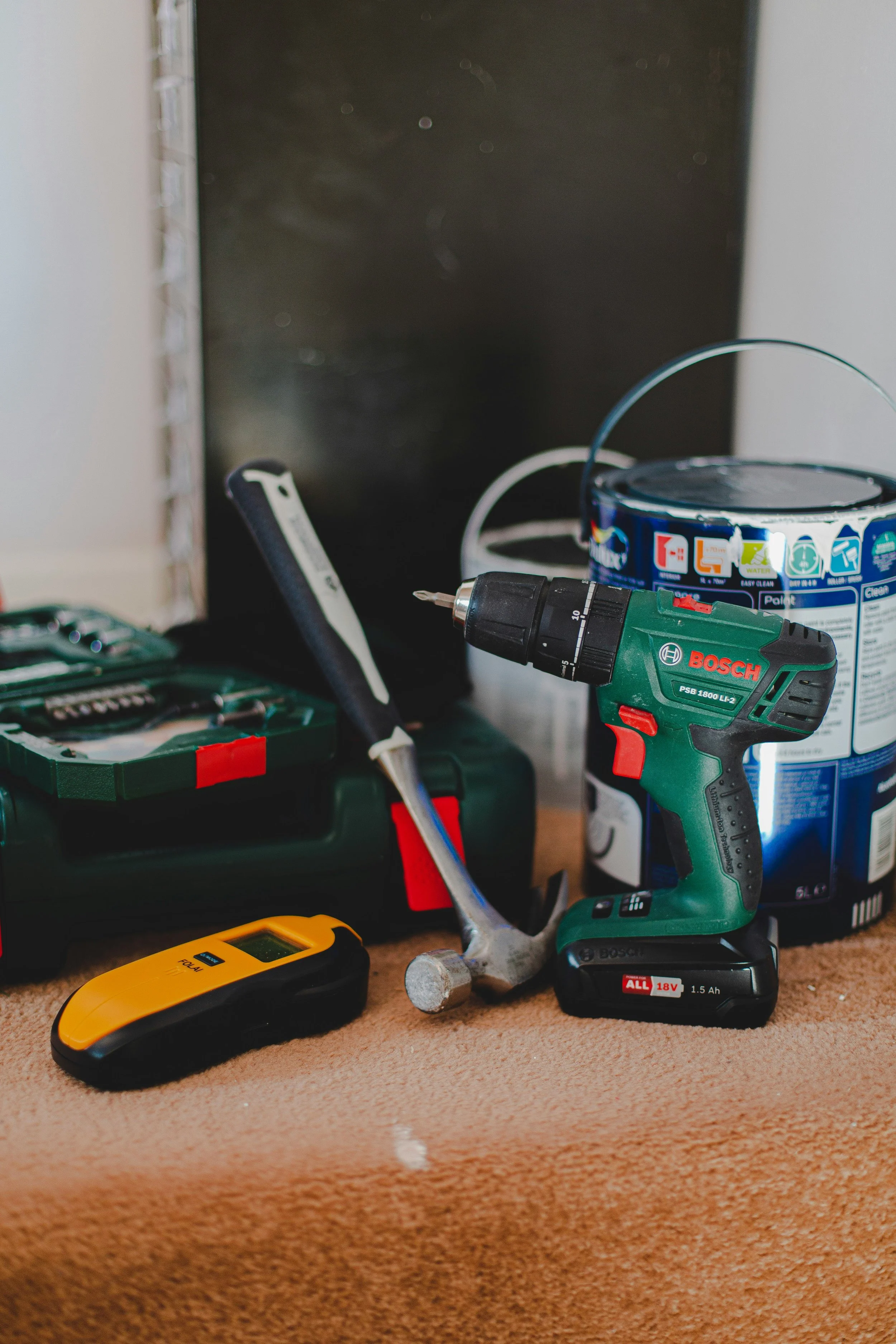 A variety of tools and supplies on a tan carpeted surface, including a Bosch cordless drill, a black and yellow laser distance meter, an open toolbox with utensils, a pen, a small yellow tape measure, and a paint can with a black lid.