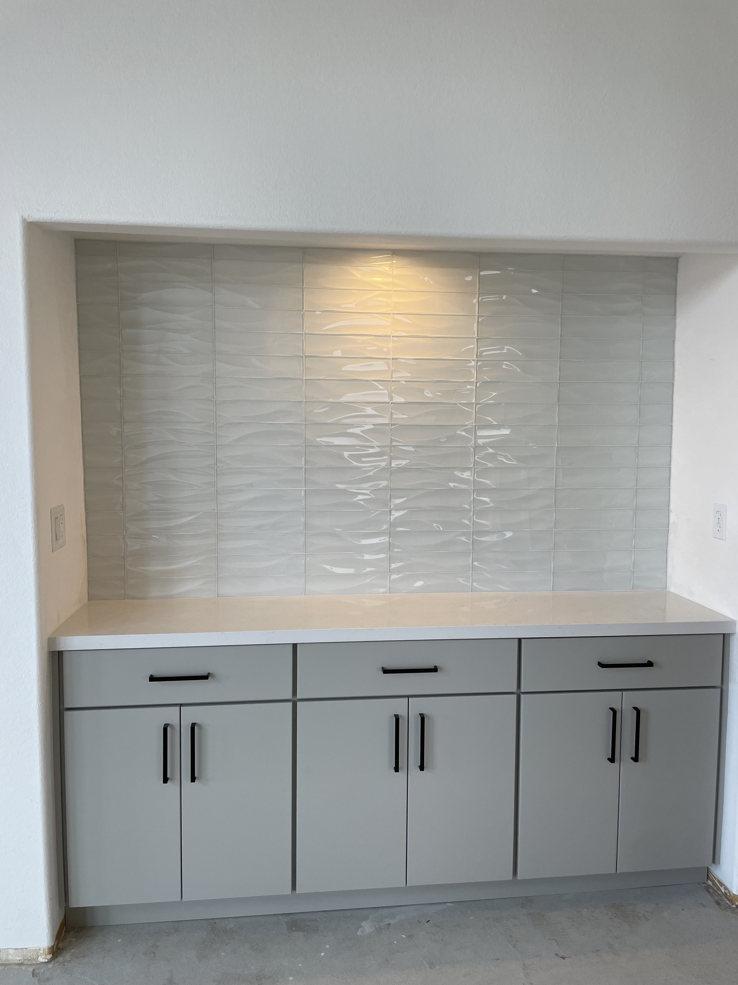 Gray kitchen cabinet with black handles beneath a counter, with a tiled backsplash featuring white rectangular tiles with a textured wave pattern, set in an alcove with white walls.