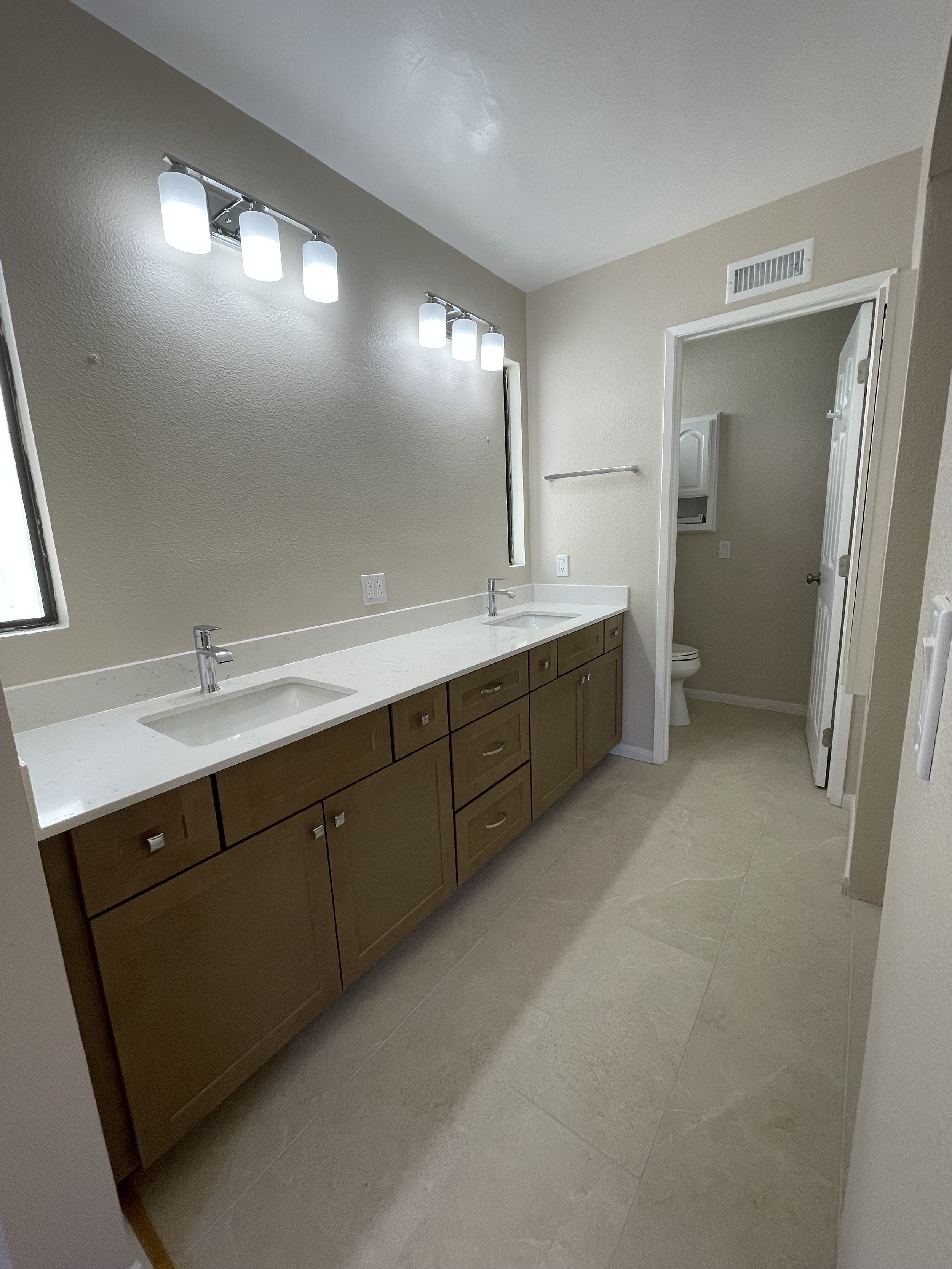 A bathroom with a double sink vanity, large mirror, and beige cabinets, with a toilet and small cabinet visible through an open door.
