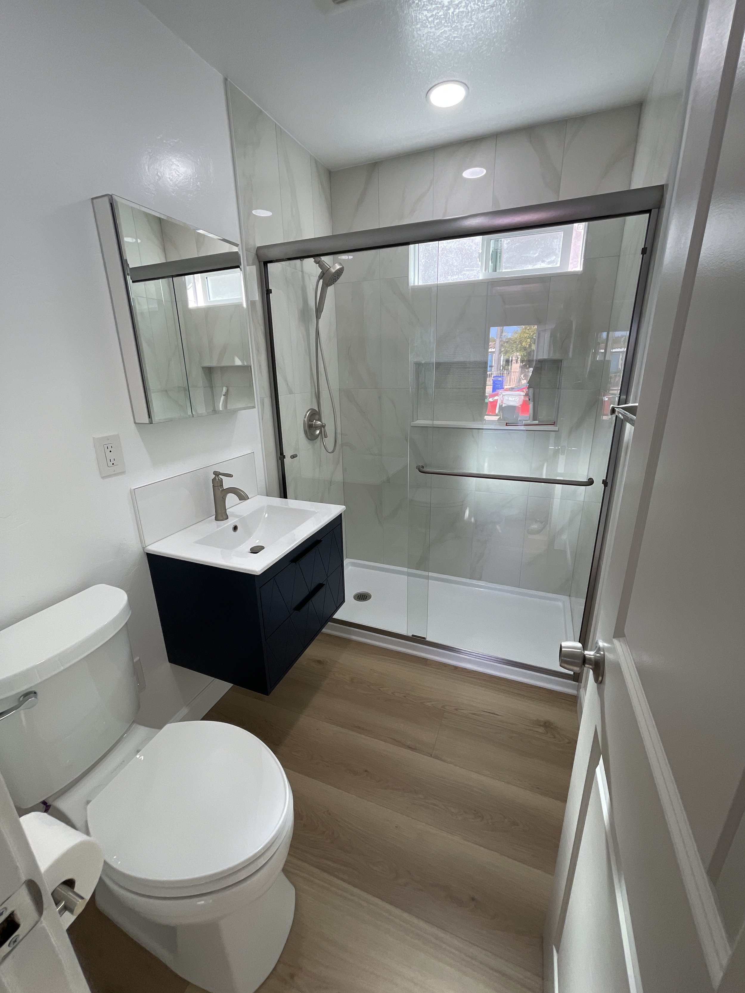 Modern bathroom with a white toilet, a vanity with a sink, a mirror cabinet, a glass shower enclosure with a sliding door, and a small window, with wood floor and recessed ceiling lights.