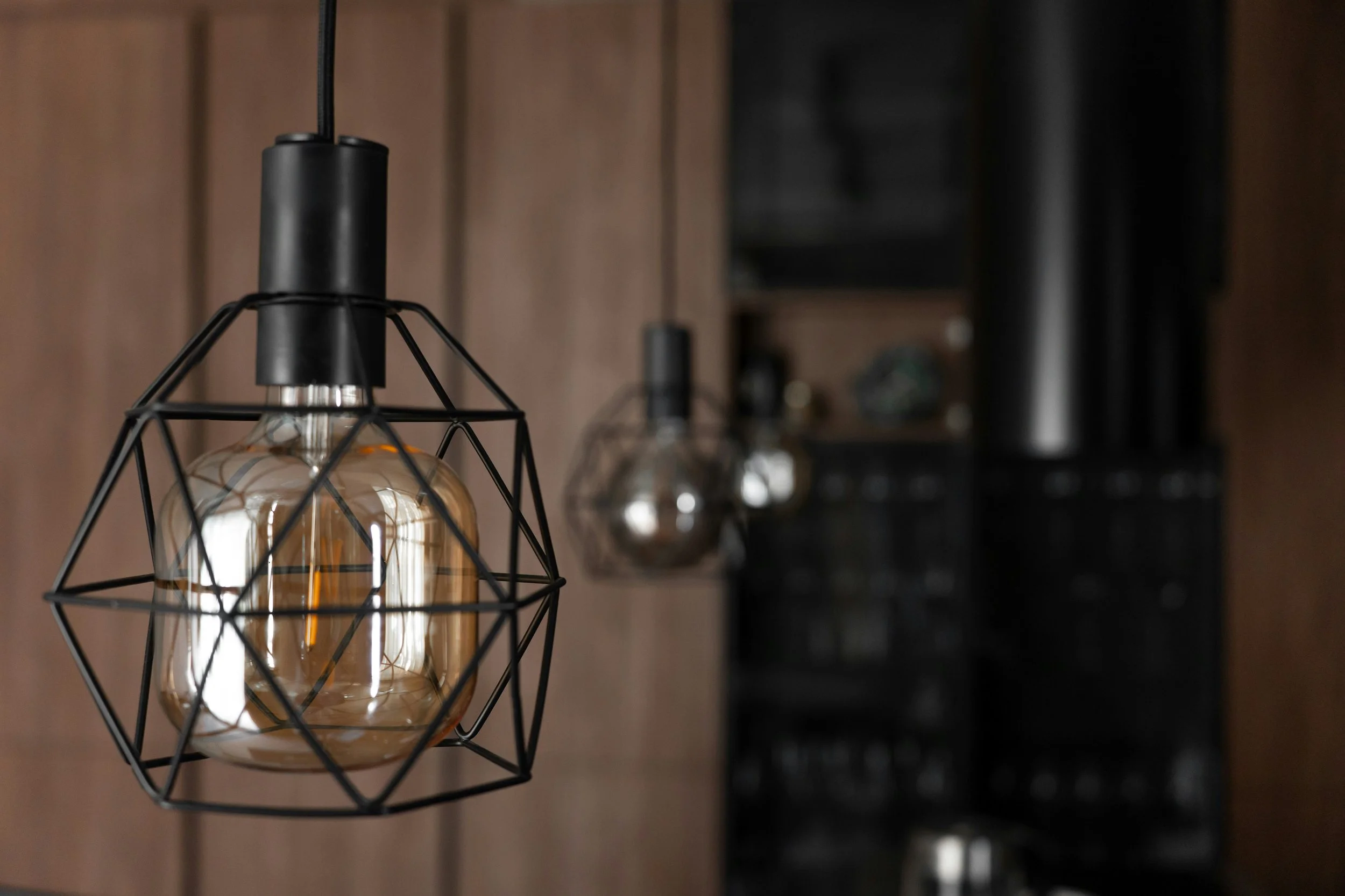 Close-up of a modern black wireframe pendant light with an Edison-style filament bulb, hanging in a room with wooden walls and a dark background.