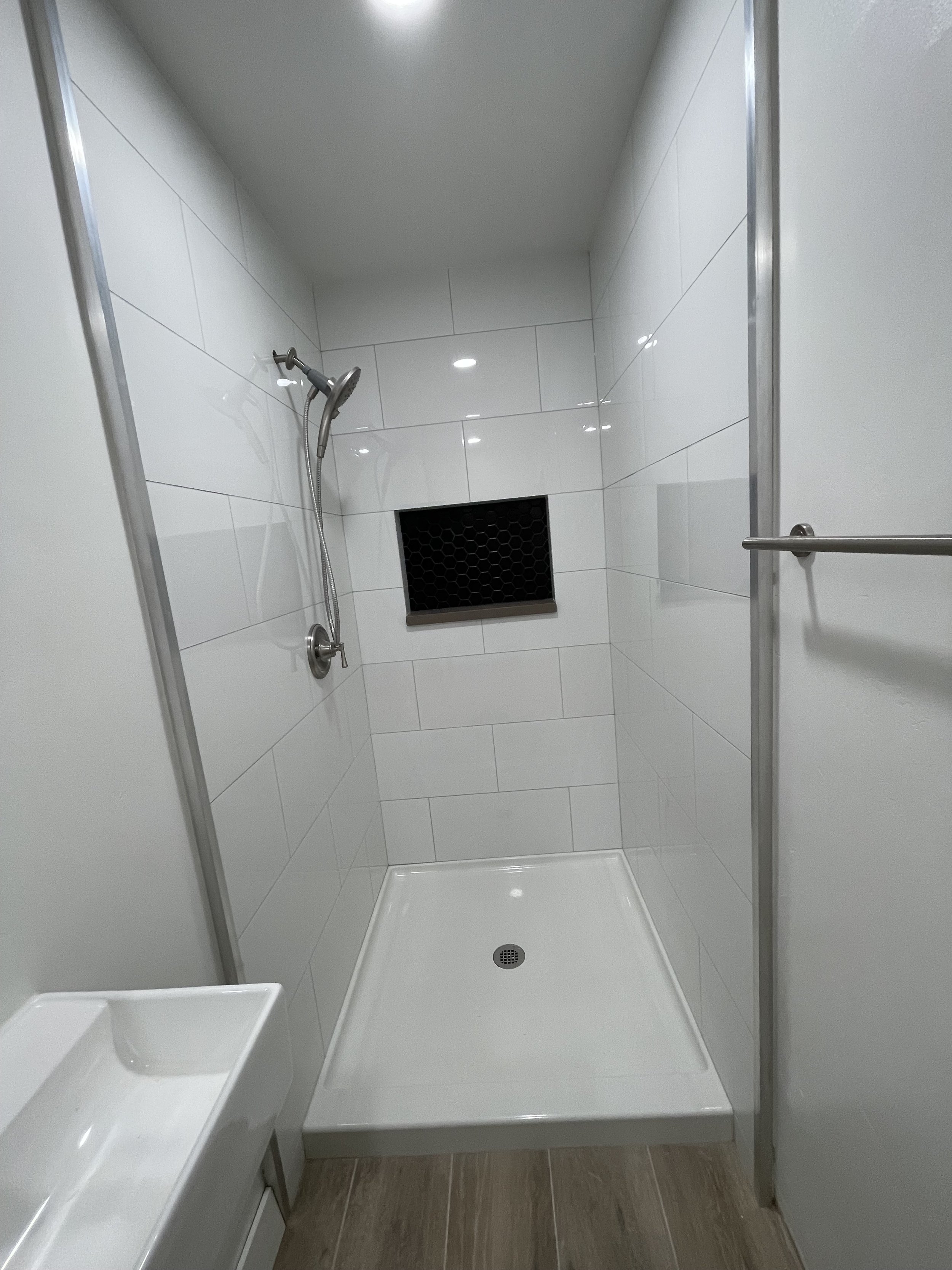 A walk-in shower with white tiled walls, a handheld shower head, a black ventilation grate, and a small corner soap dish. The shower has a glass door and a metal railing on one side.