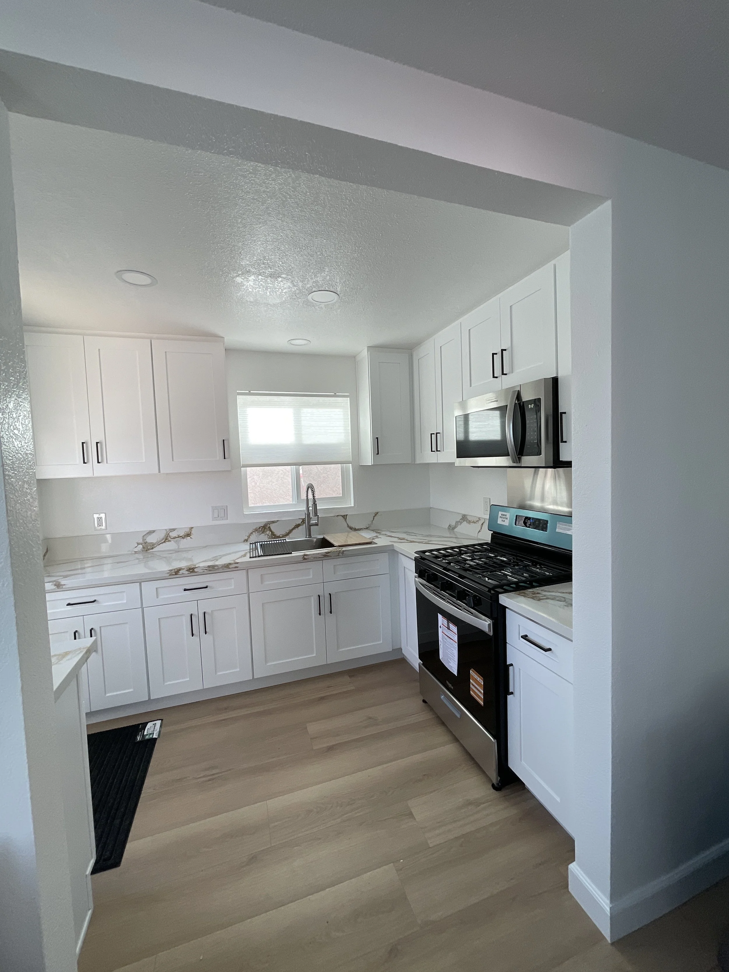 Modern kitchen with white cabinets, a marble countertop, stainless steel appliances, a window with blinds, and light wood flooring.