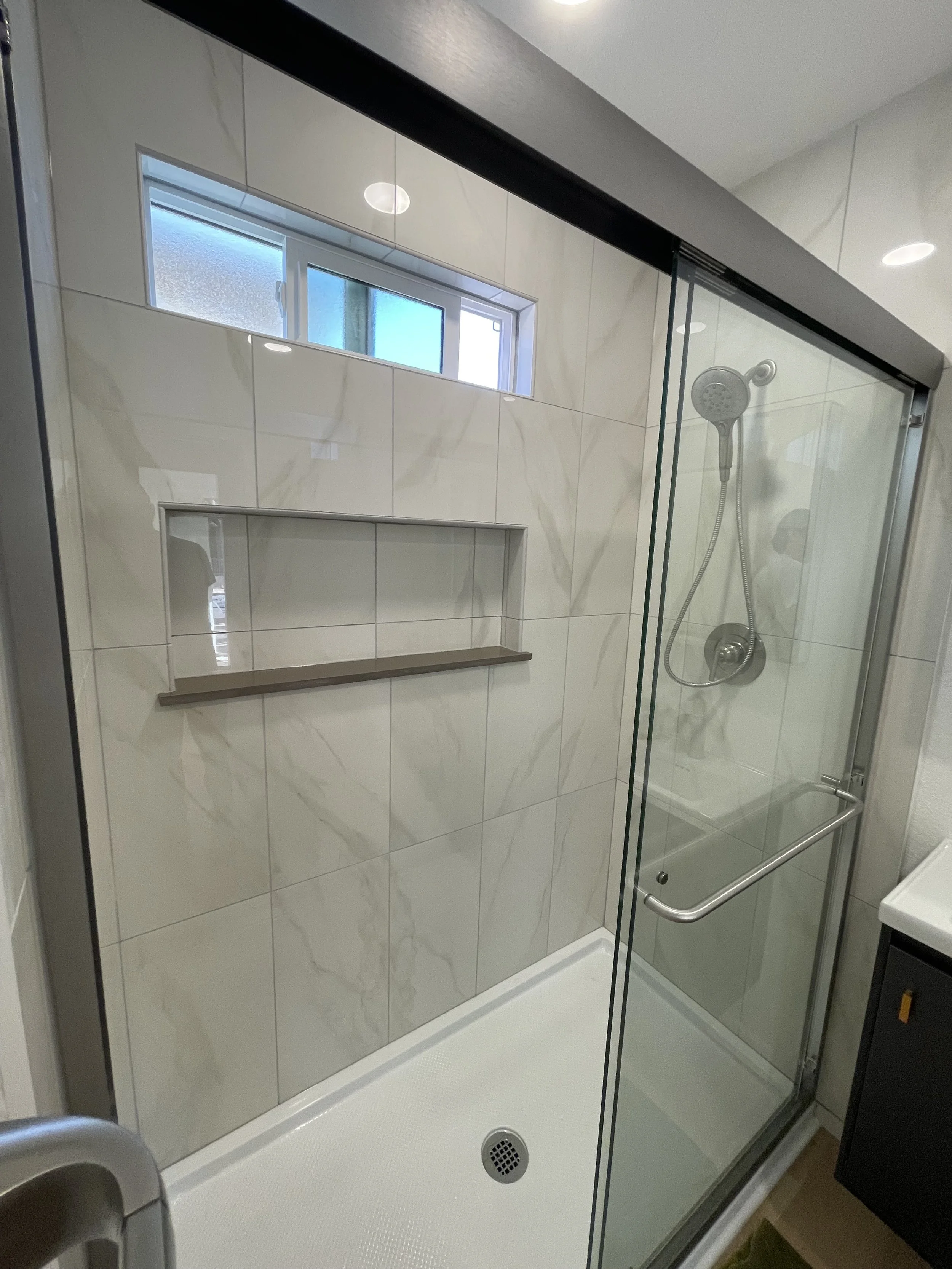 A modern bathroom shower with white marbled tiles, a small horizontal frosted window, a corner niche with a wooden shelf, a showerhead with a detachable hose, and a glass door with metal hinges and handle.