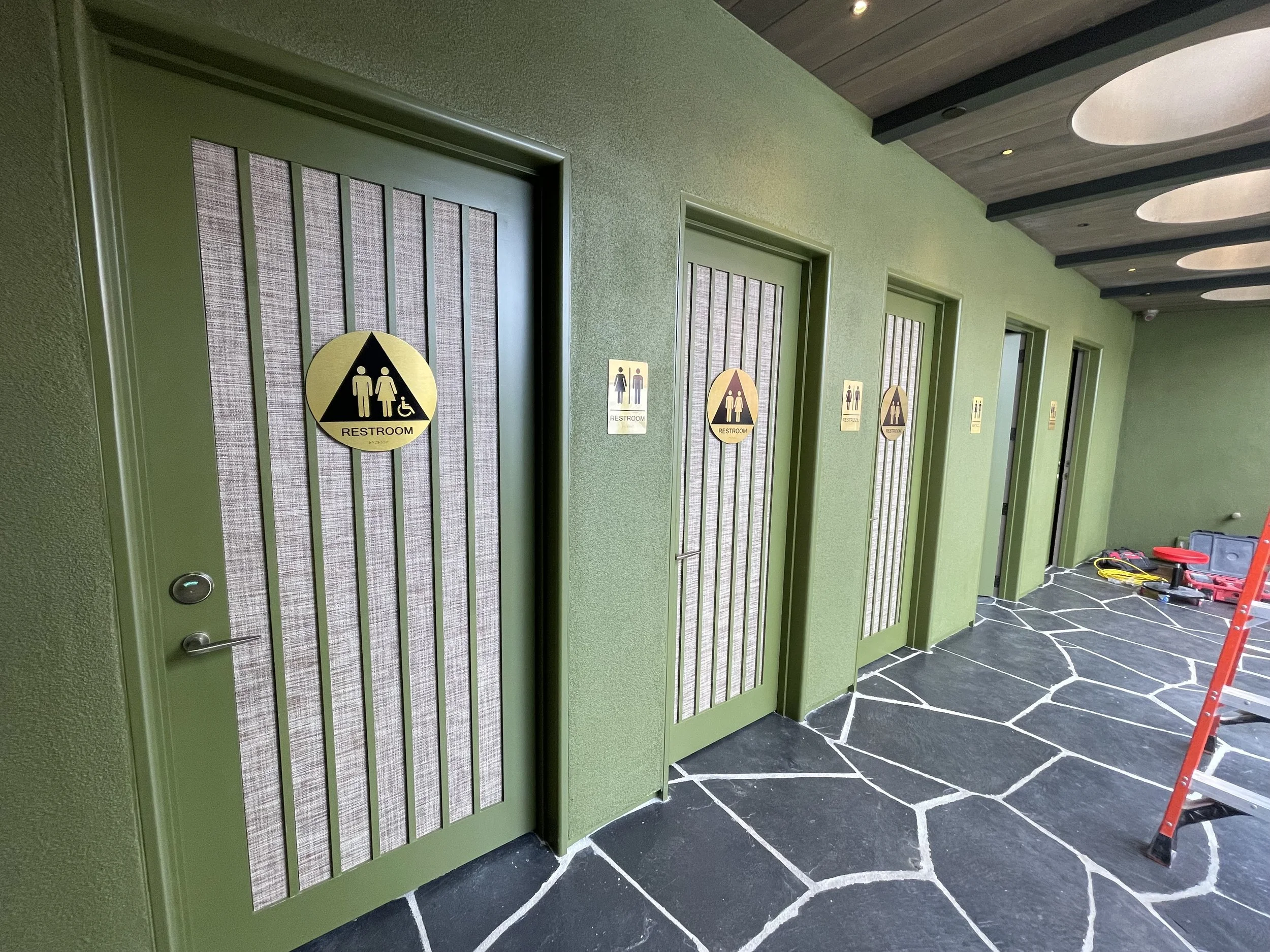 A row of green bathroom stall doors with signs indicating restrooms, some with wheelchair accessibility symbols, in a modern building corridor with black tile flooring featuring white grout lines, and a toolbox and ladder on the floor.