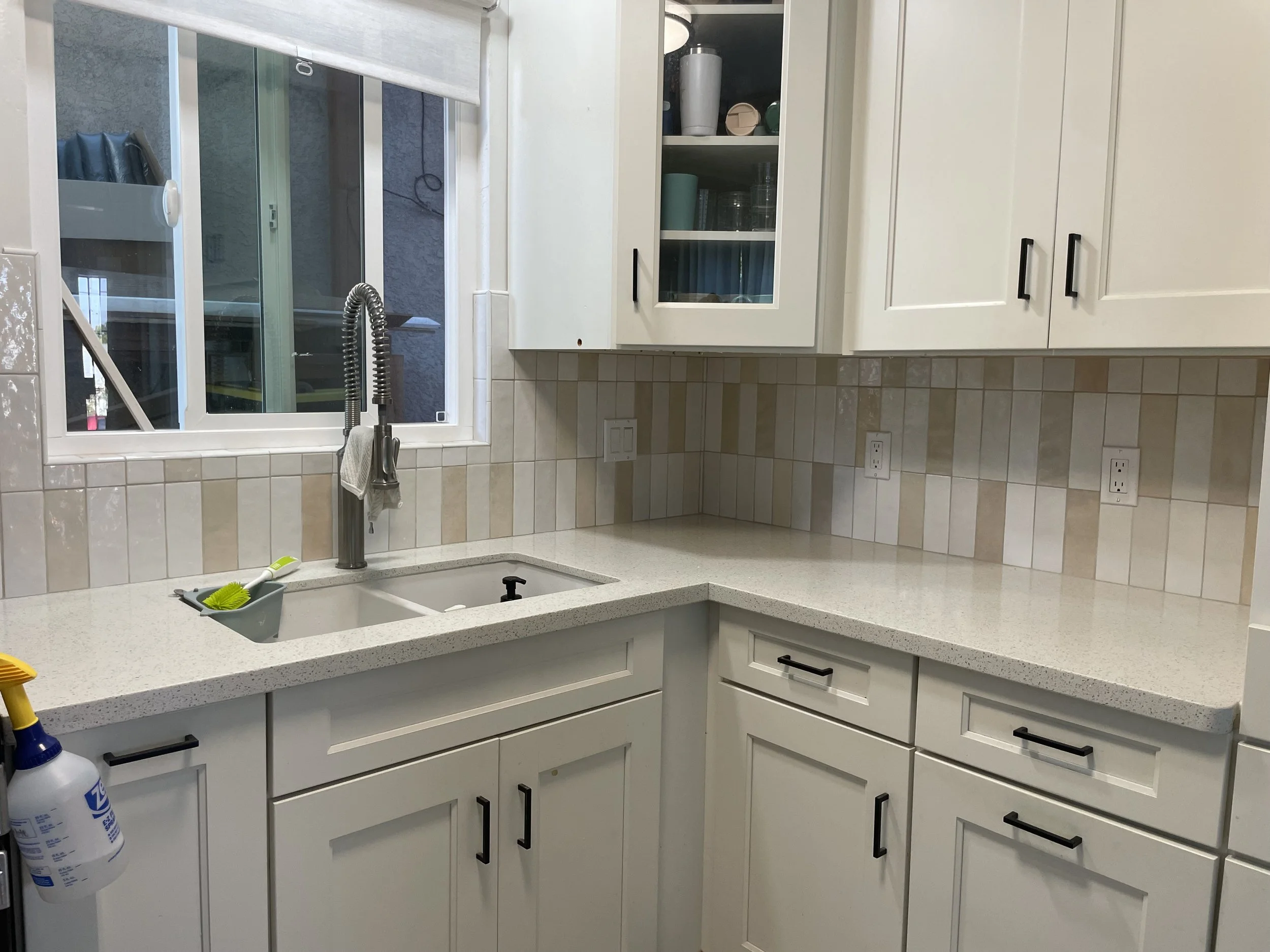 Kitchen with white cabinets and beige tiled backsplash, stainless steel sink, window above the sink, and cleaning supplies on the counter.