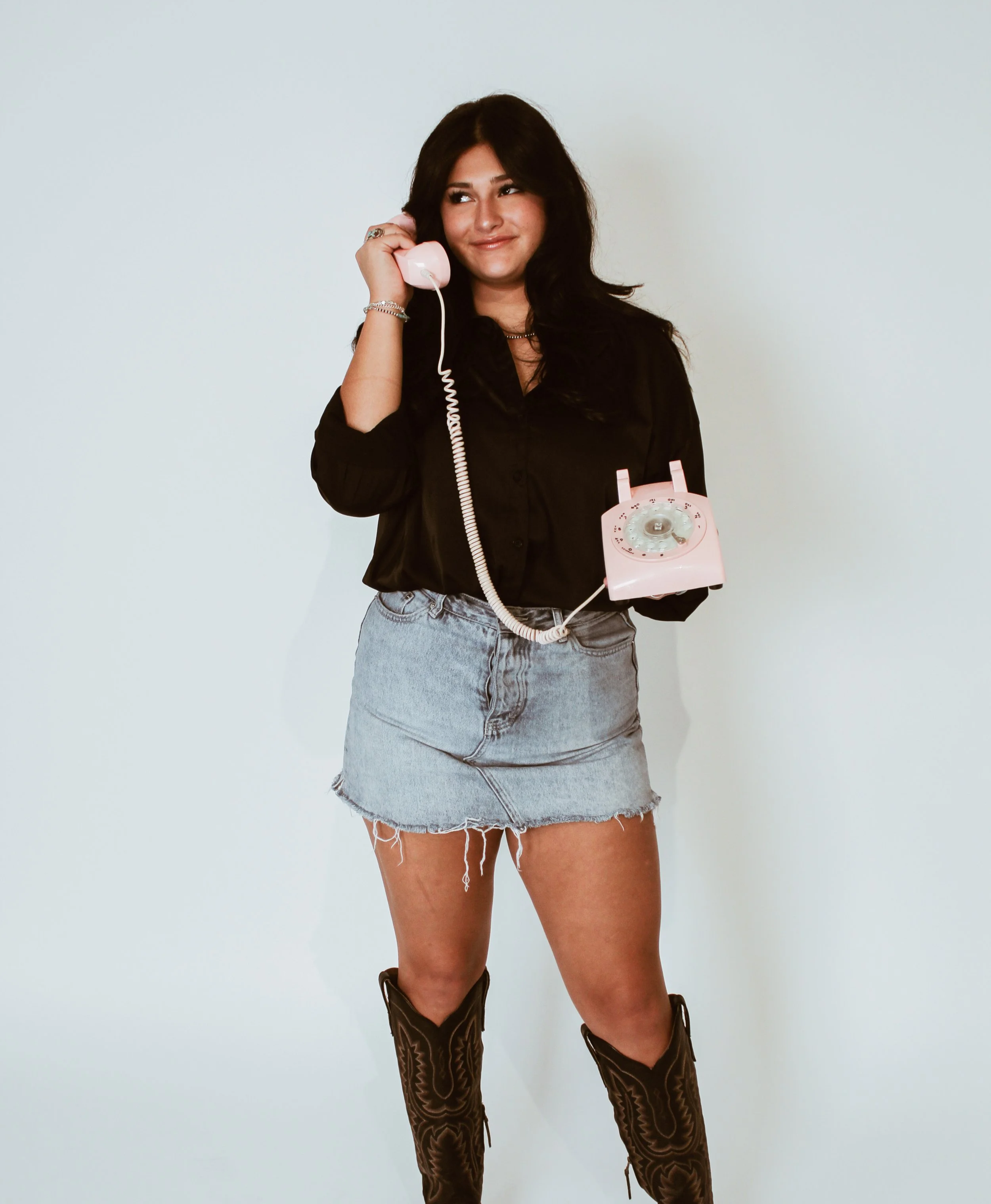 Young woman with dark hair and wearing cowboy boots, denim skirt, and black shirt, holding a pink rotary phone to her ear, standing against a plain light background.
