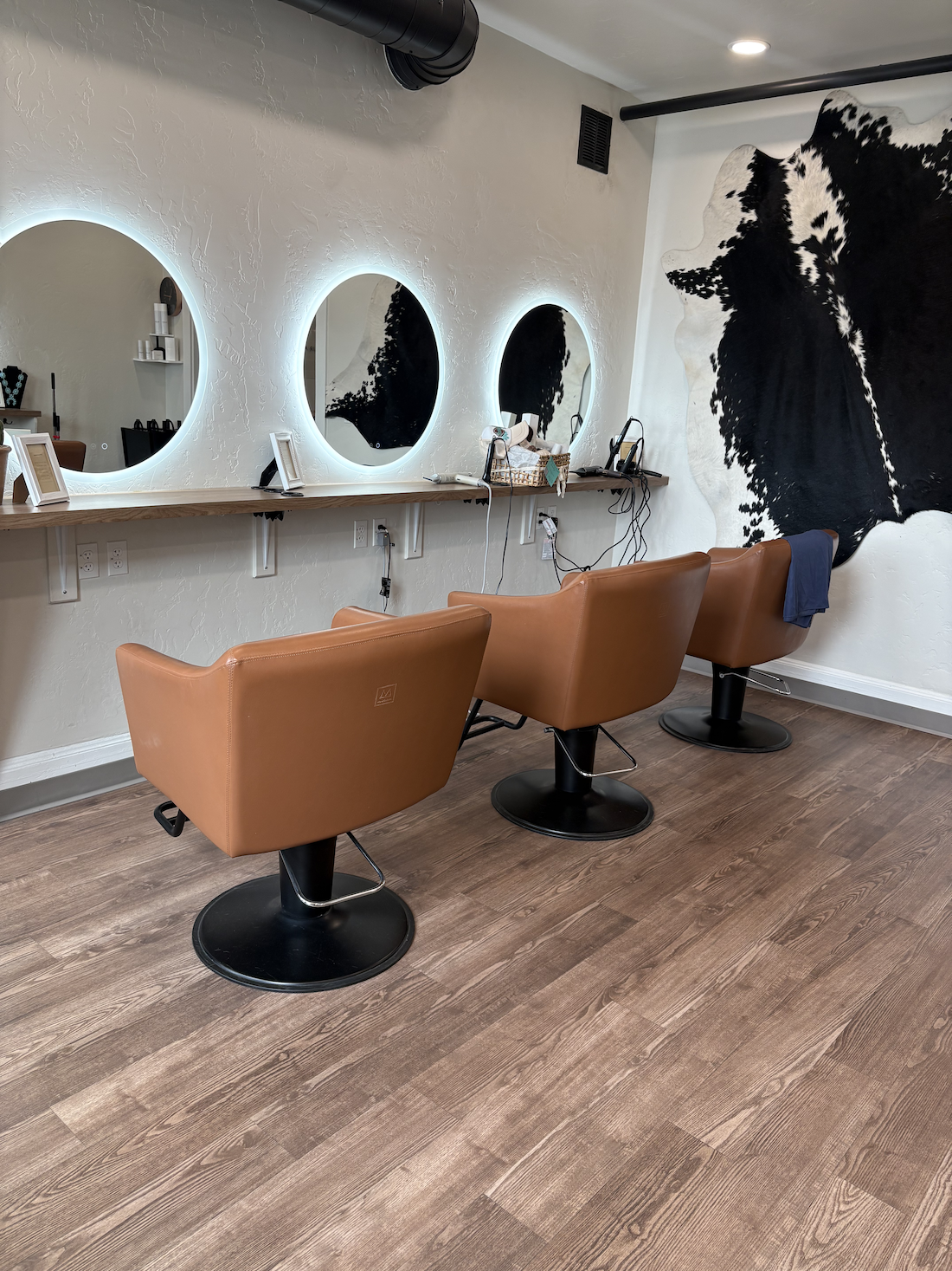 A hair salon station with three chairs, three illuminated mirrors, a large cowhide wall art piece, and wooden flooring.