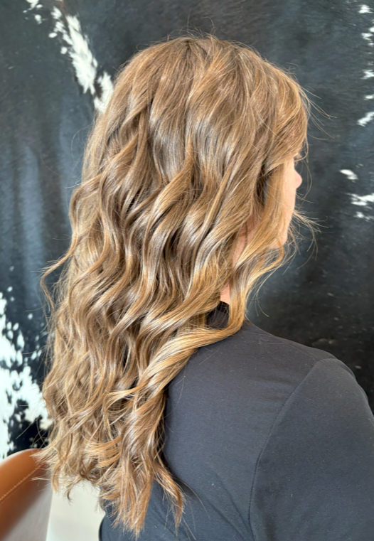 Side view of a woman with long, wavy, light brown hair wearing a black top, indoors against a dark background with white streaks.