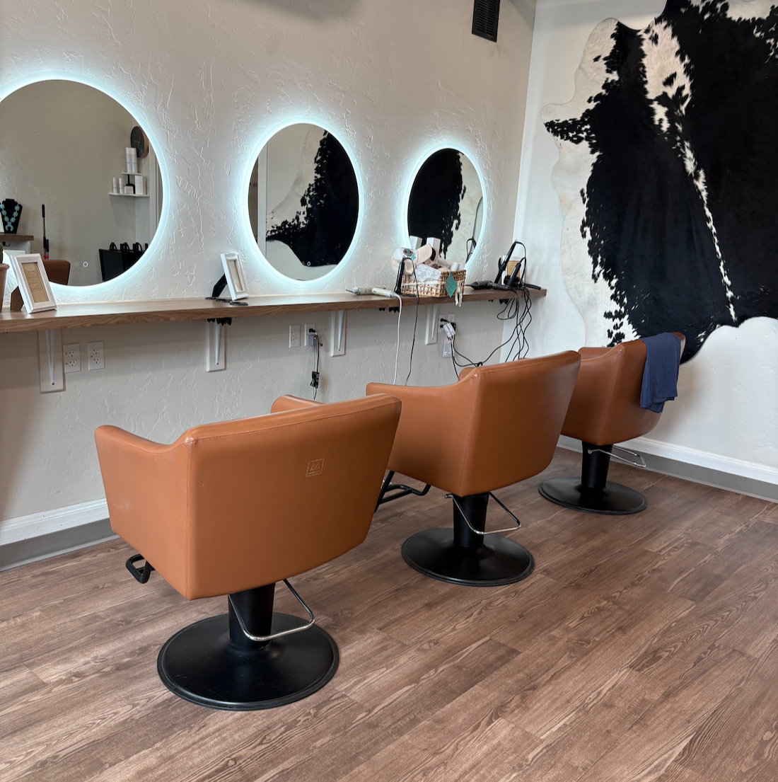 Salon with three brown styling chairs in front of a wall with three illuminated mirrors, and a large black and white illustration of a woman's profile on the wall.