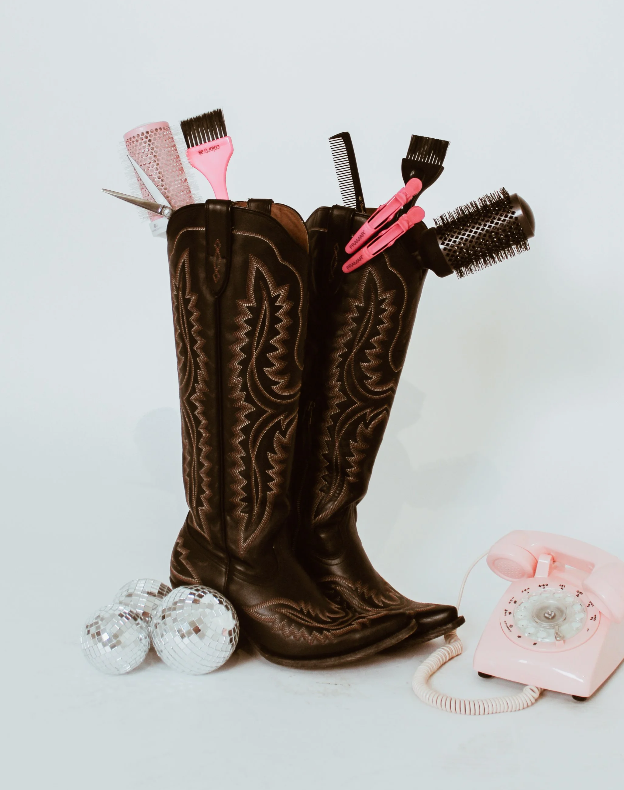 A vintage black leather cowboy boot filled with pink hair styling tools and brushes, surrounded by disco balls and a pink rotary telephone on a plain white background.