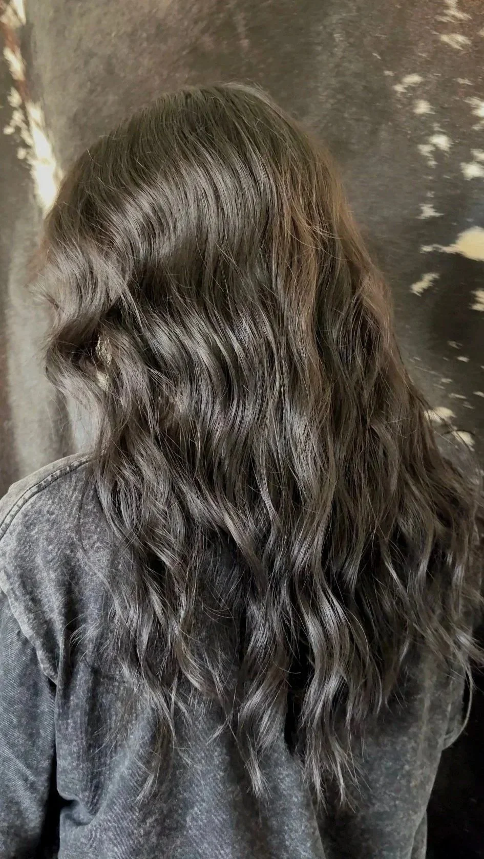 A person with long, wavy, silver-gray hair seen from behind, wearing a gray top, standing against a dark textured wall.