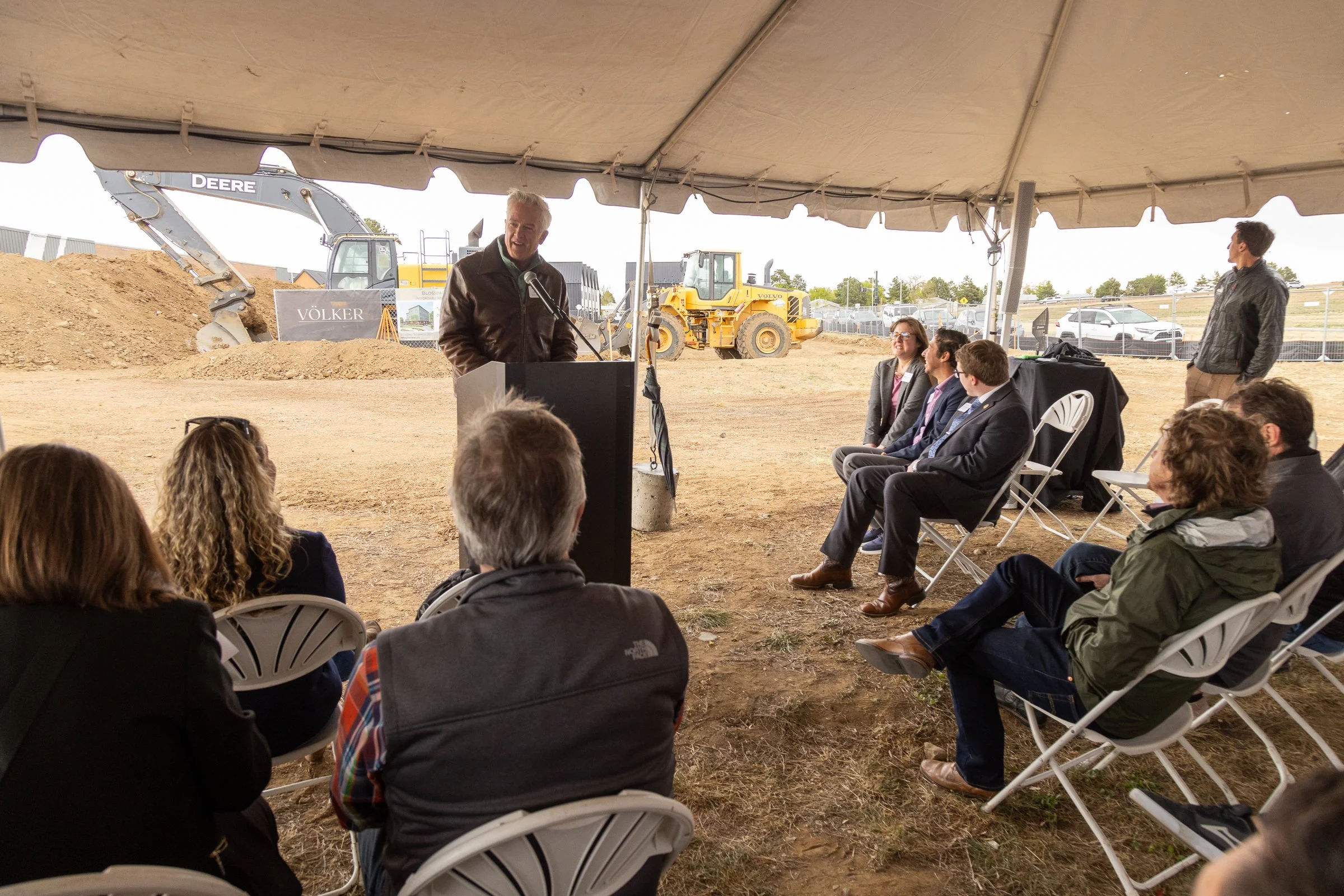 A man is giving a speech at an outdoor construction site under a large tent, with construction equipment like a backhoe and a wheel loader visible in the background. Several people are seated and listening, while others stand nearby.