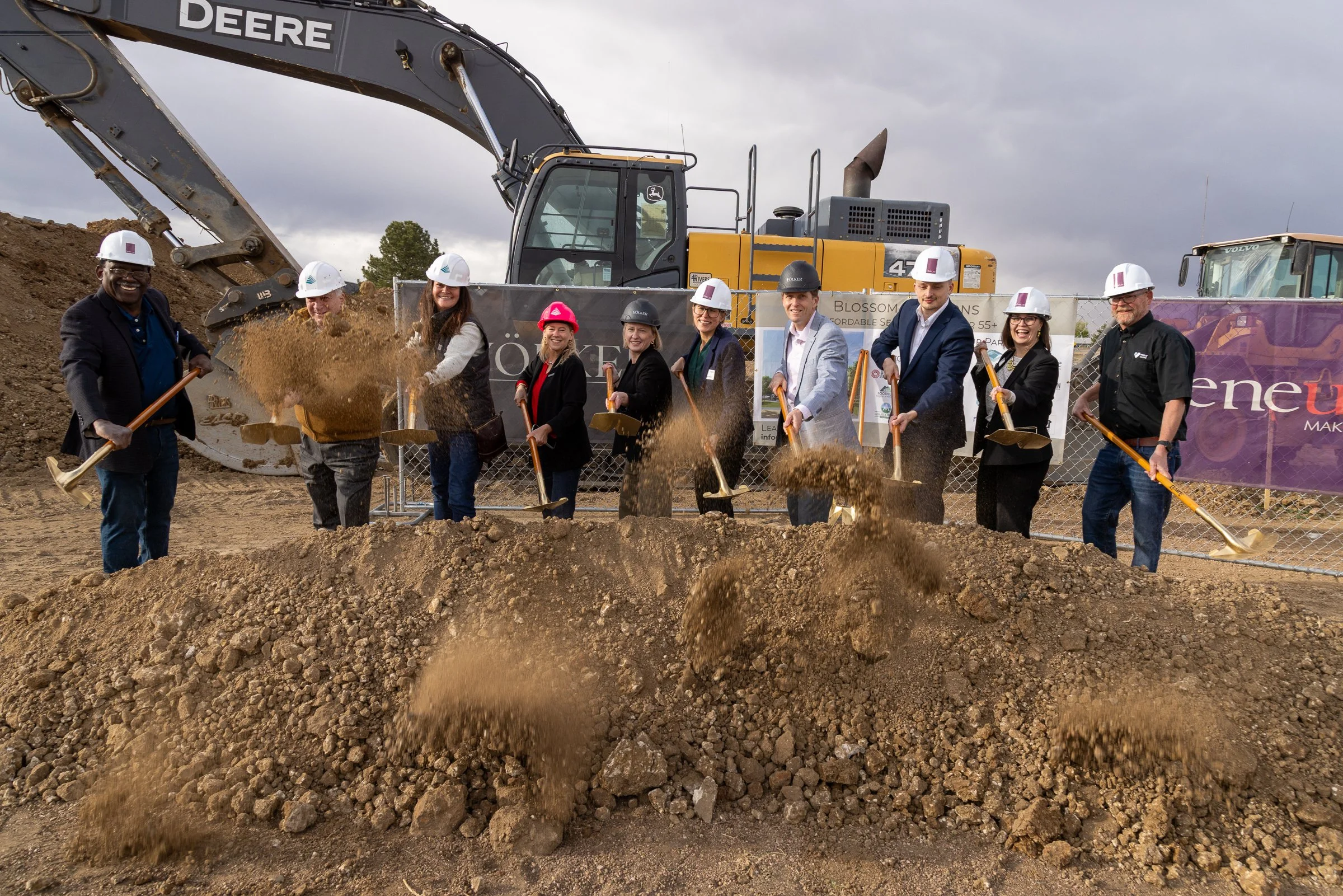 Group of people in hard hats planting a tree at a construction site with heavy machinery and a fence in the background
