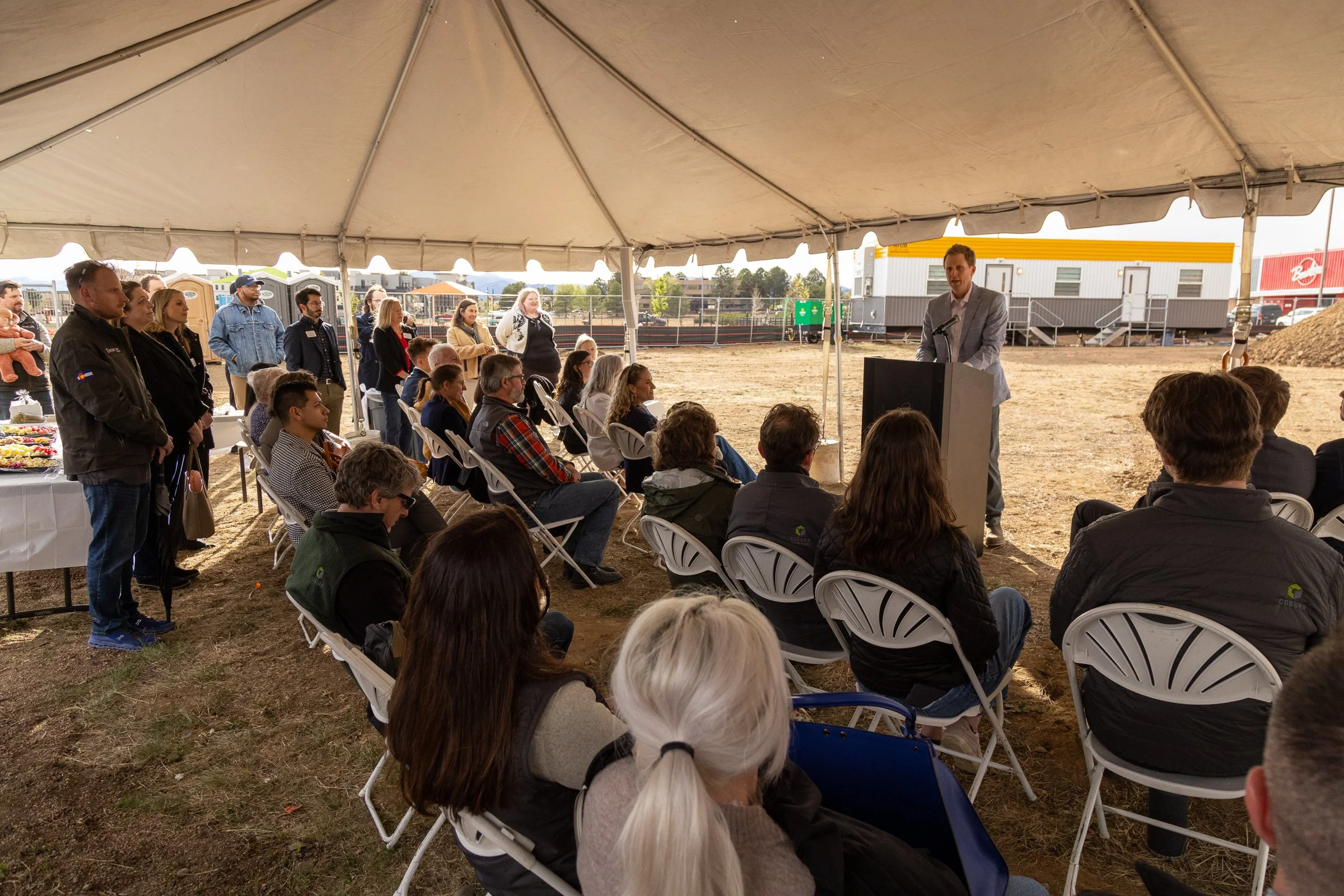 A group of people seated and standing under a large tent, attending a presentation outdoors. A man in a gray suit is speaking at a podium, facing the audience. The background includes portable toilets, portable buildings, and outdoor scenery.