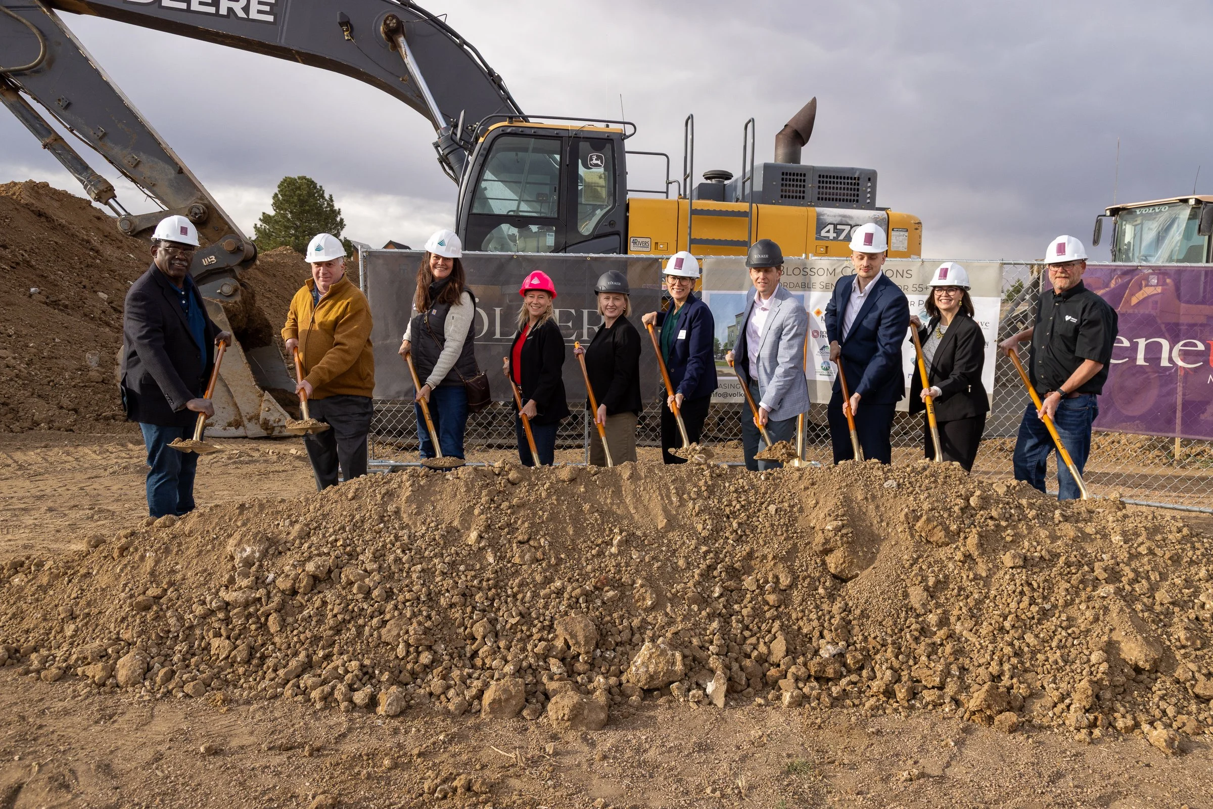 A group of people wearing safety helmets and holding shovels standing in front of construction equipment at a groundbreaking ceremony. There are large excavators behind them and a fence with banners.