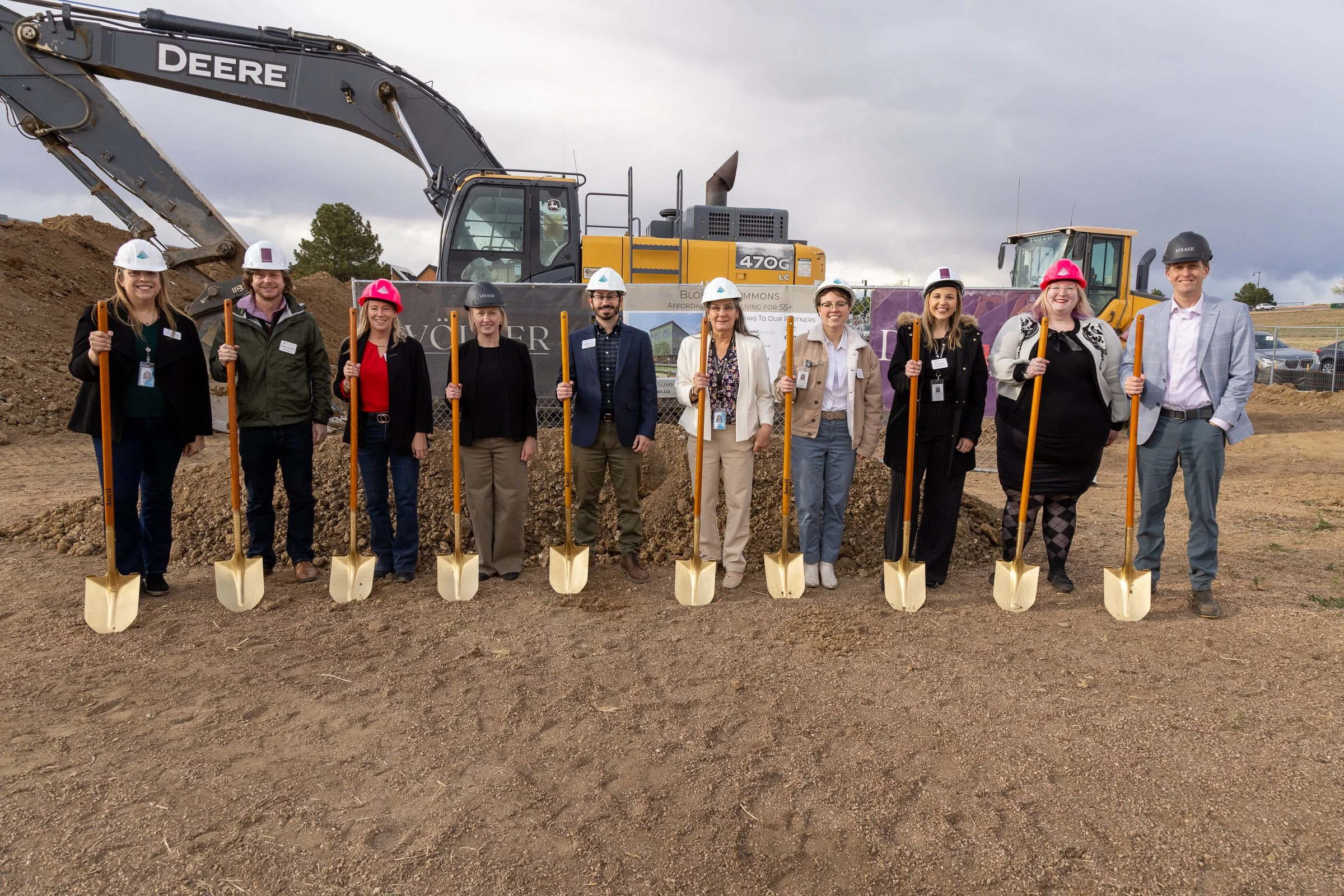 Group of nine people standing with shovels in front of construction equipment at a construction site, some wearing safety helmets and badges, posing for photo with cloudy sky in background.