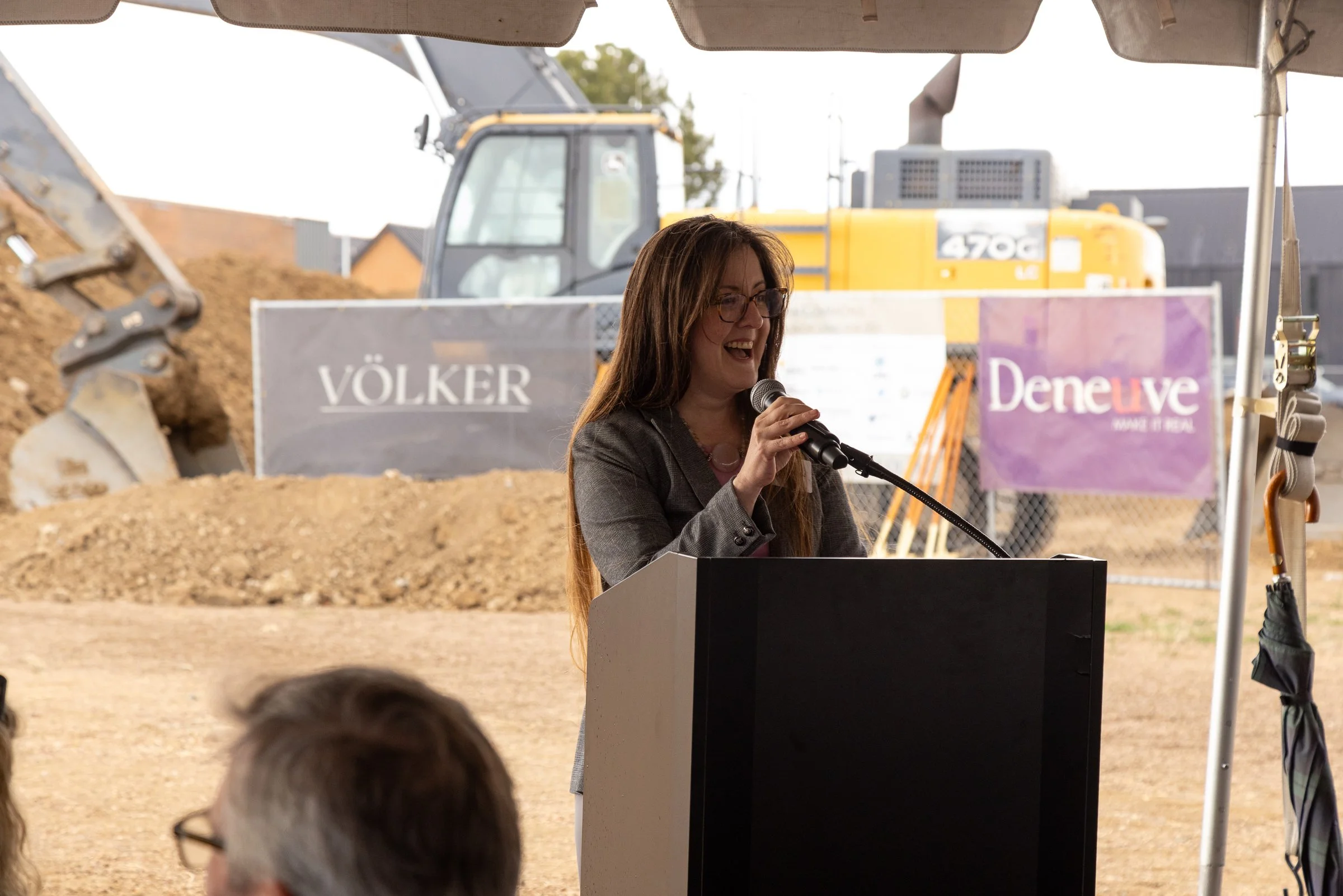 A woman with long brown hair and glasses, smiling and speaking into a microphone at a podium outdoors during daytime. Behind her, there is construction equipment, including a yellow excavator, and a large sign with the word "VÖLKER" on it. The settin