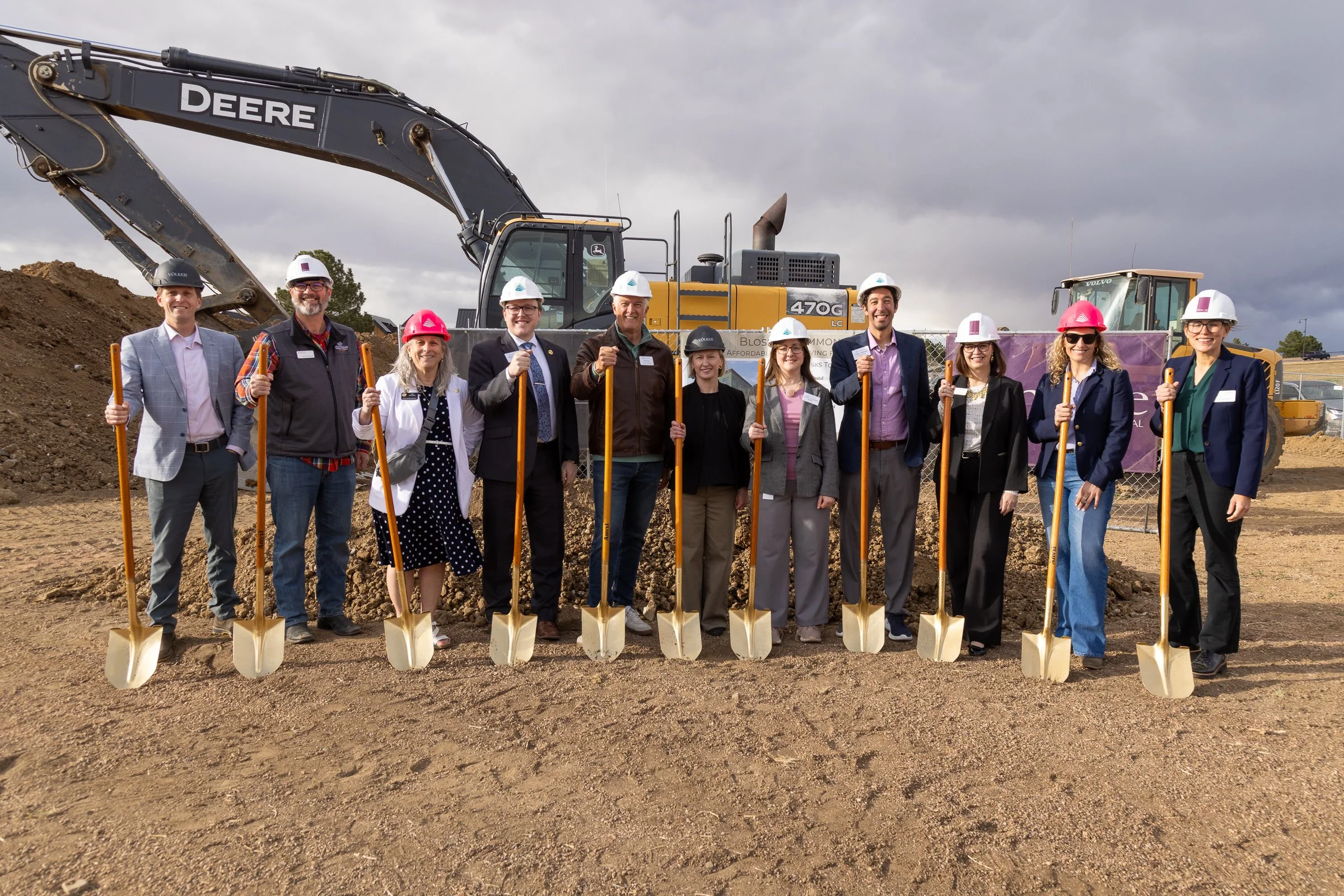 Group of people in hard hats and business attire standing with shovels at a construction site, with excavators and dirt in the background.