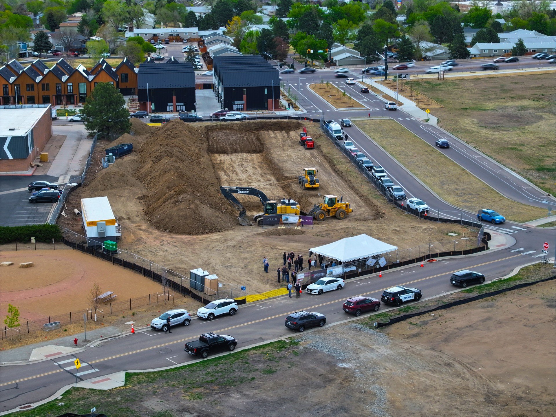 A construction site in a residential area with a large dirt excavation, construction machinery, and a temporary tent. There are parked cars and a small group of people gathered near the site, with some viewing from a sidewalk and others near the cons